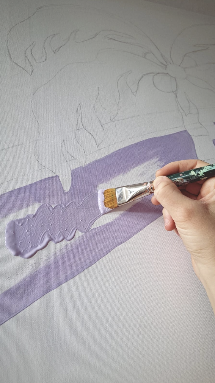 A person is applying light purple paint with a wide brush onto a canvas with an outline of a flower in the background.