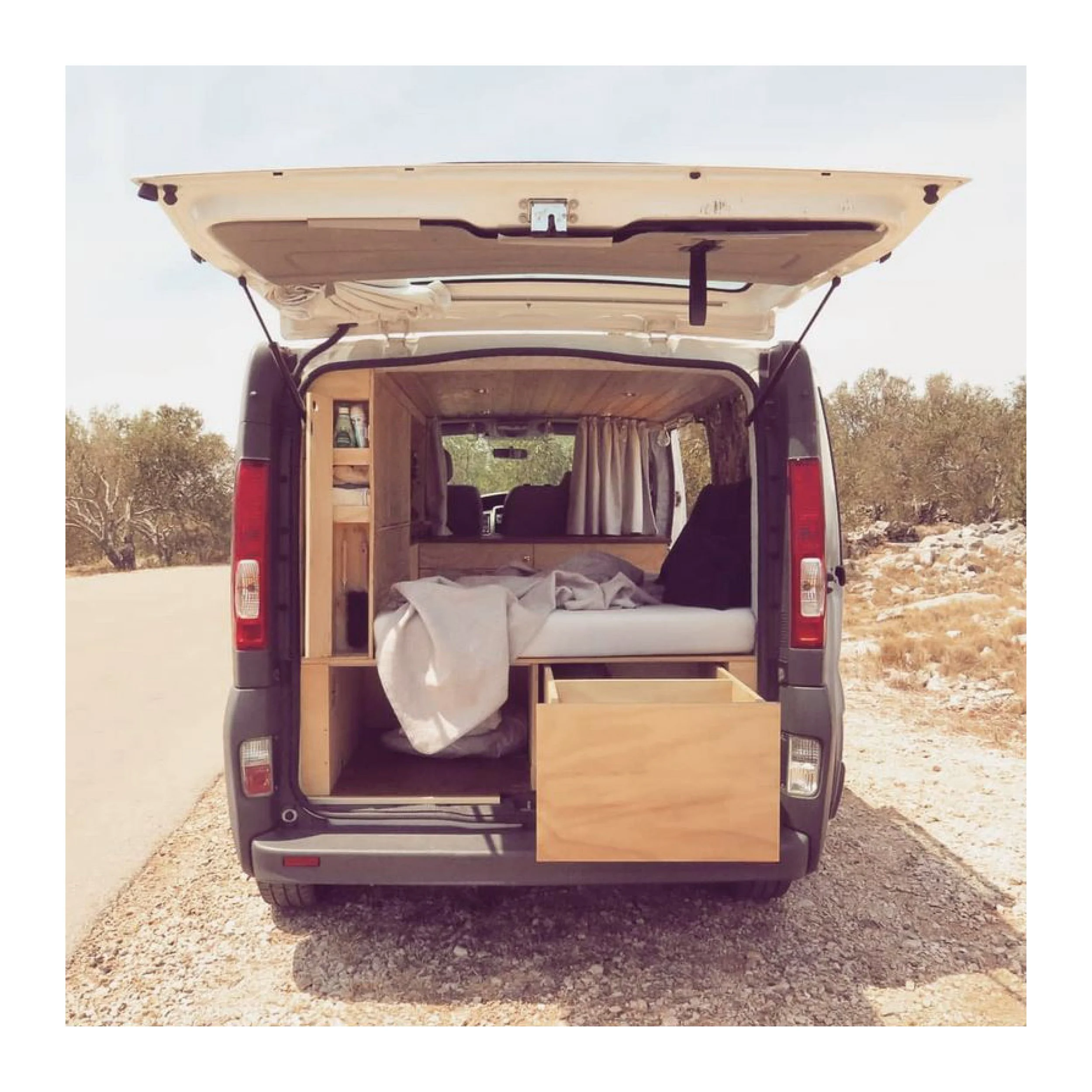 The rear of a camper van with an open roof hatch, showing a built-in bed, curtains, storage shelves, and wooden furniture inside, parked on a dirt road in a desert-like landscape.