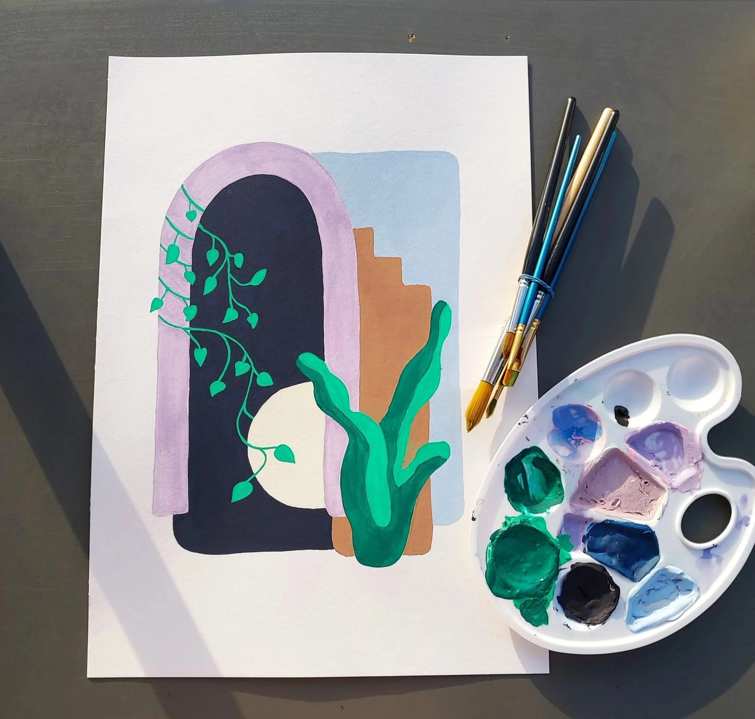 A watercolor painting of a house with an arched door, purple trim, green plants, a moon, and abstract shapes, placed on a table with painting brushes and a palette with various paint colors.