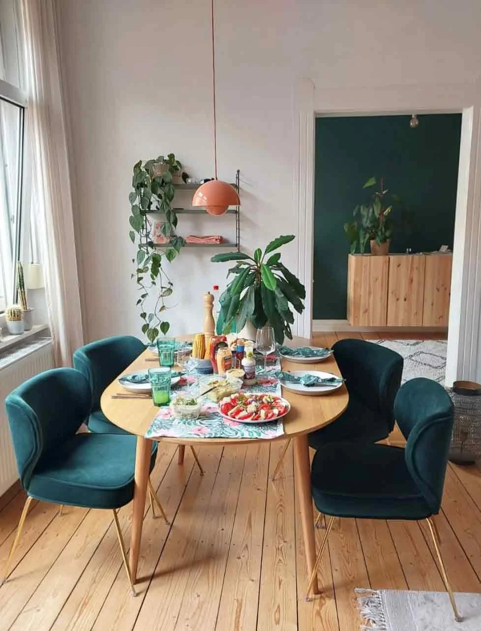 A dining room with a wooden table set for a meal, surrounded by four dark green upholstered chairs with gold legs. The table has a colorful table runner, plates, glasses, and food including a pizza. There are green plants on the table and in the room, with a window on the left letting in natural light, and a wall with shelves and hanging plants on the left side of the background.