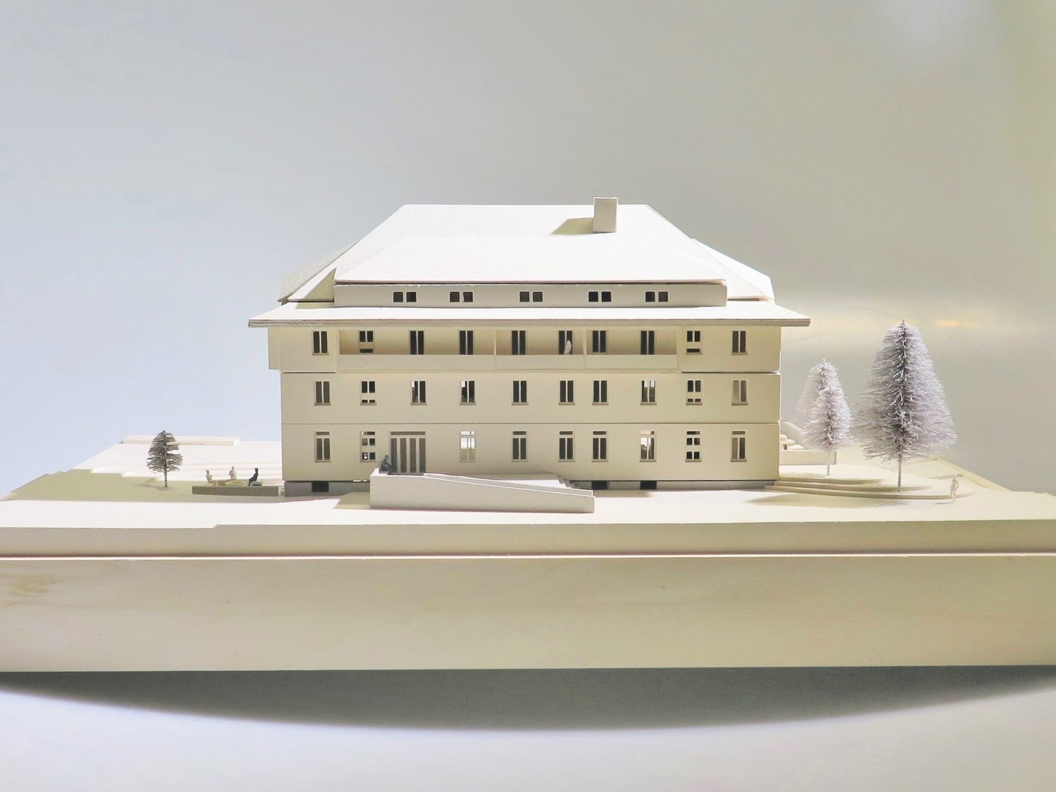 A white architectural model of a large multi-story building with a pitched roof, surrounded by small trees and miniature figures on a white platform.