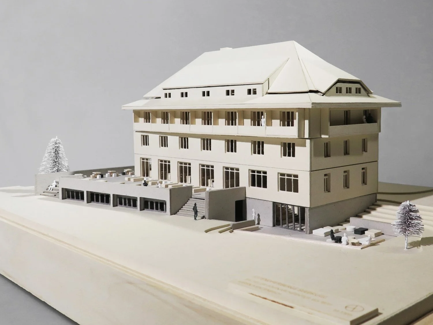 A detailed architectural model of a multi-story building with exterior balconies, stairs, and outdoor seating areas, set on a wooden base with miniature trees and figurines.