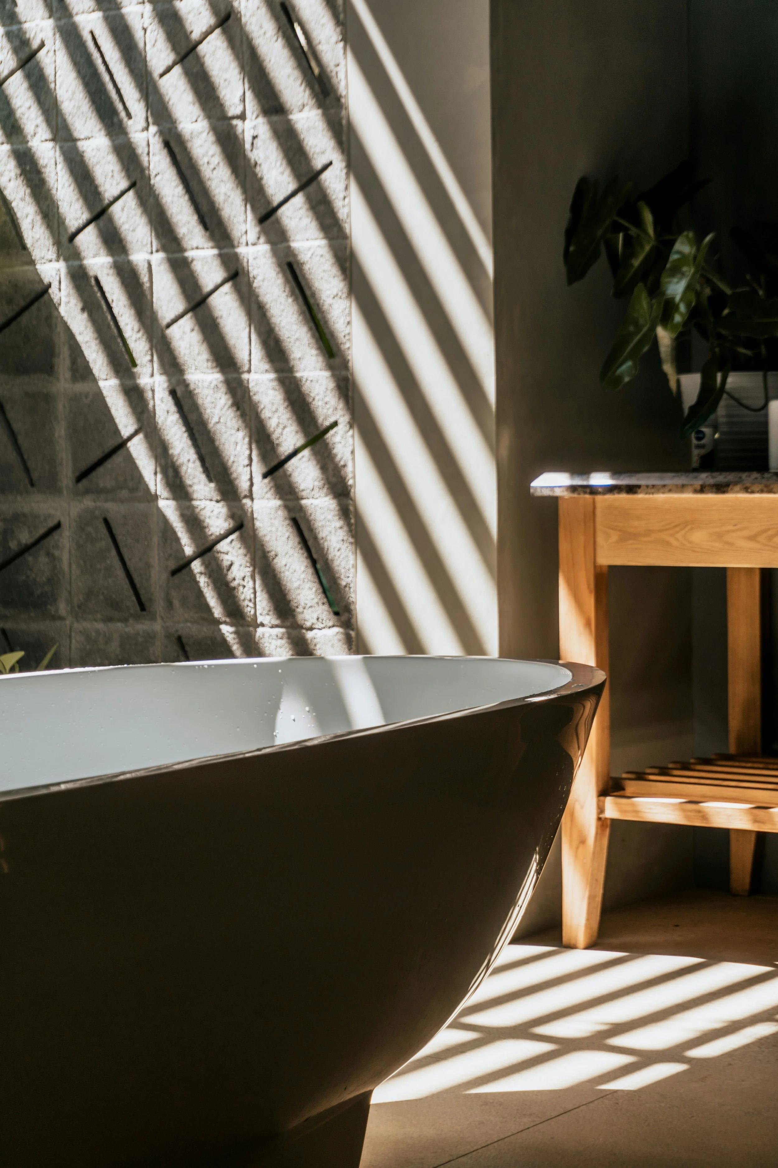 A modern bathroom with a white bathtub, casting shadows of window blinds onto the wall and floor, with a wooden shelf and a green plant nearby.
