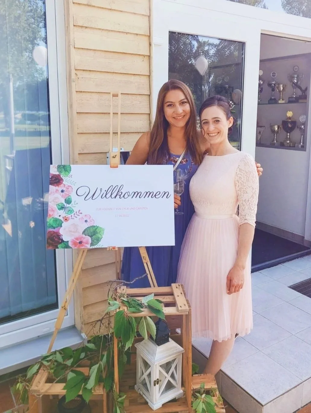 Two women standing at a wedding reception, smiling, holding drinks, and posing next to a welcoming sign that says 'Willkommen'. The sign has floral decorations and text in German. The women are dressed in elegant dresses, one in blue and the other in white, with a backdrop of a glass door and trophy display case inside the venue.