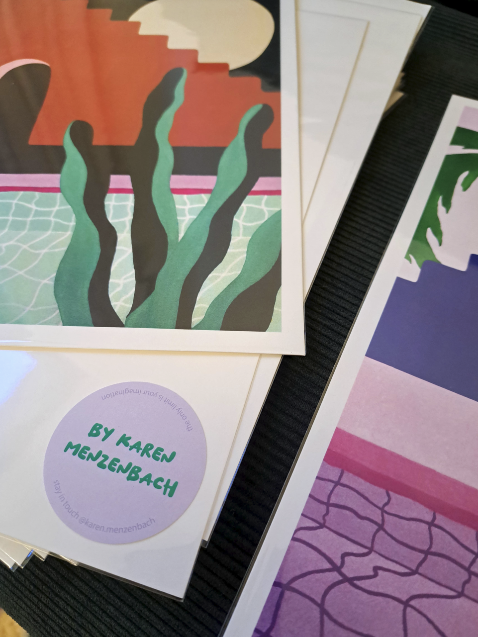 Close-up of a collection of colorful printed art posters and a circular sticker with green text reading "BY KAREN MENZENBACH," on a black textured surface.