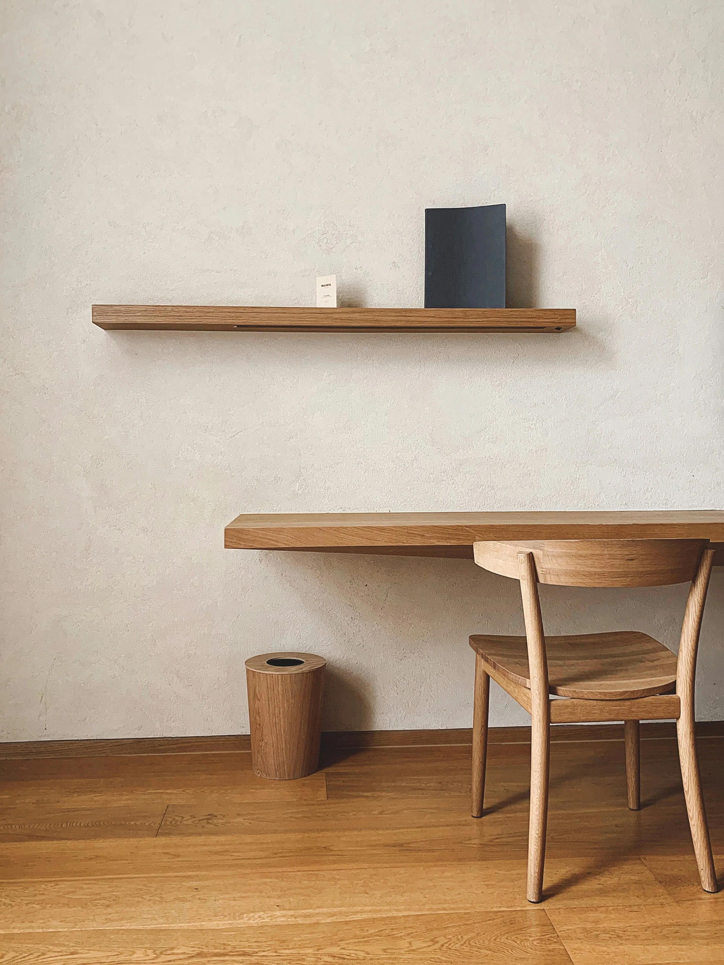 Minimalist interior with a wooden desk, wooden chair, a small wooden trash bin, and two floating wooden shelves on a textured white wall.