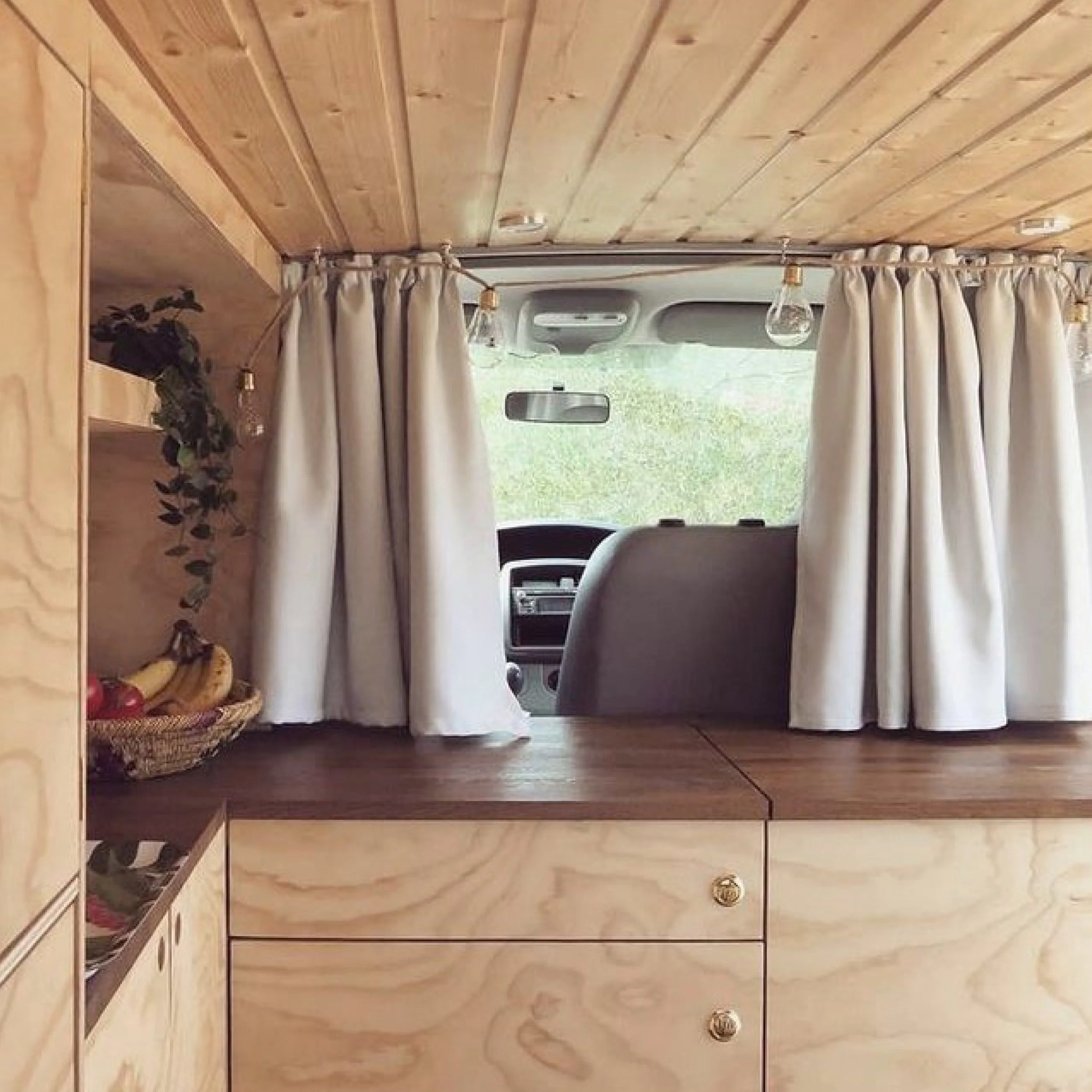 Interior view of a converted camper van with a wooden cabinet and cream curtains, showing the front dash and windshield with outside greenery.