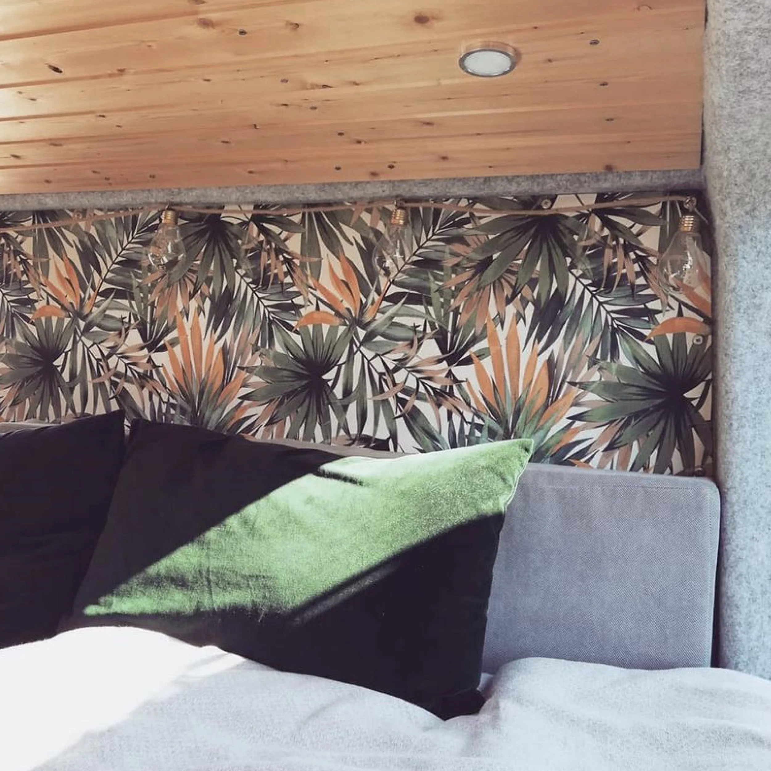 A bedroom with a bed, black and green velvet pillows, a patterned wallpaper with tropical leaves, a wooden ceiling, and hanging light bulbs.
