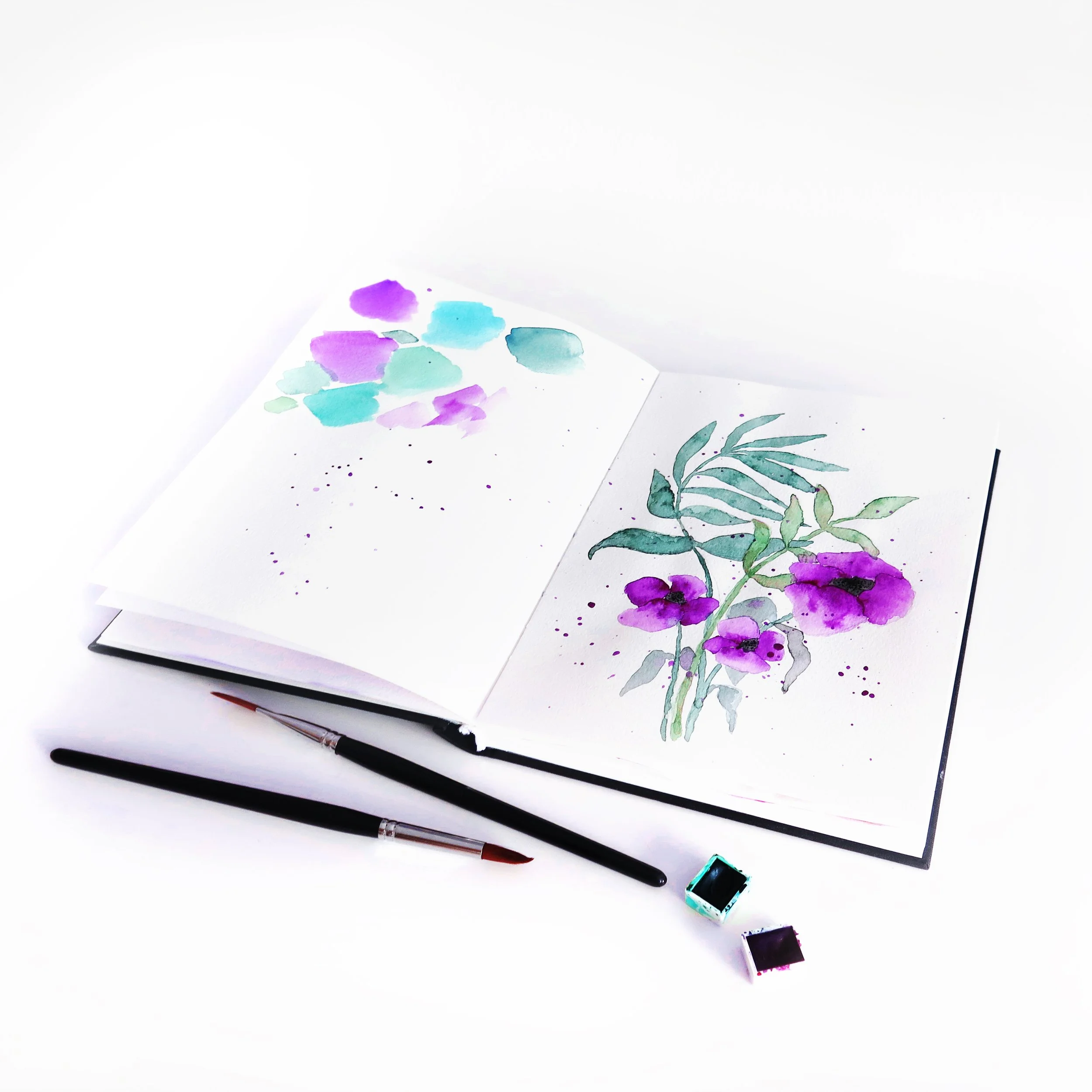 Open sketchbook with watercolor flower illustrations, watercolor paints, paintbrushes, and ink on a white background.