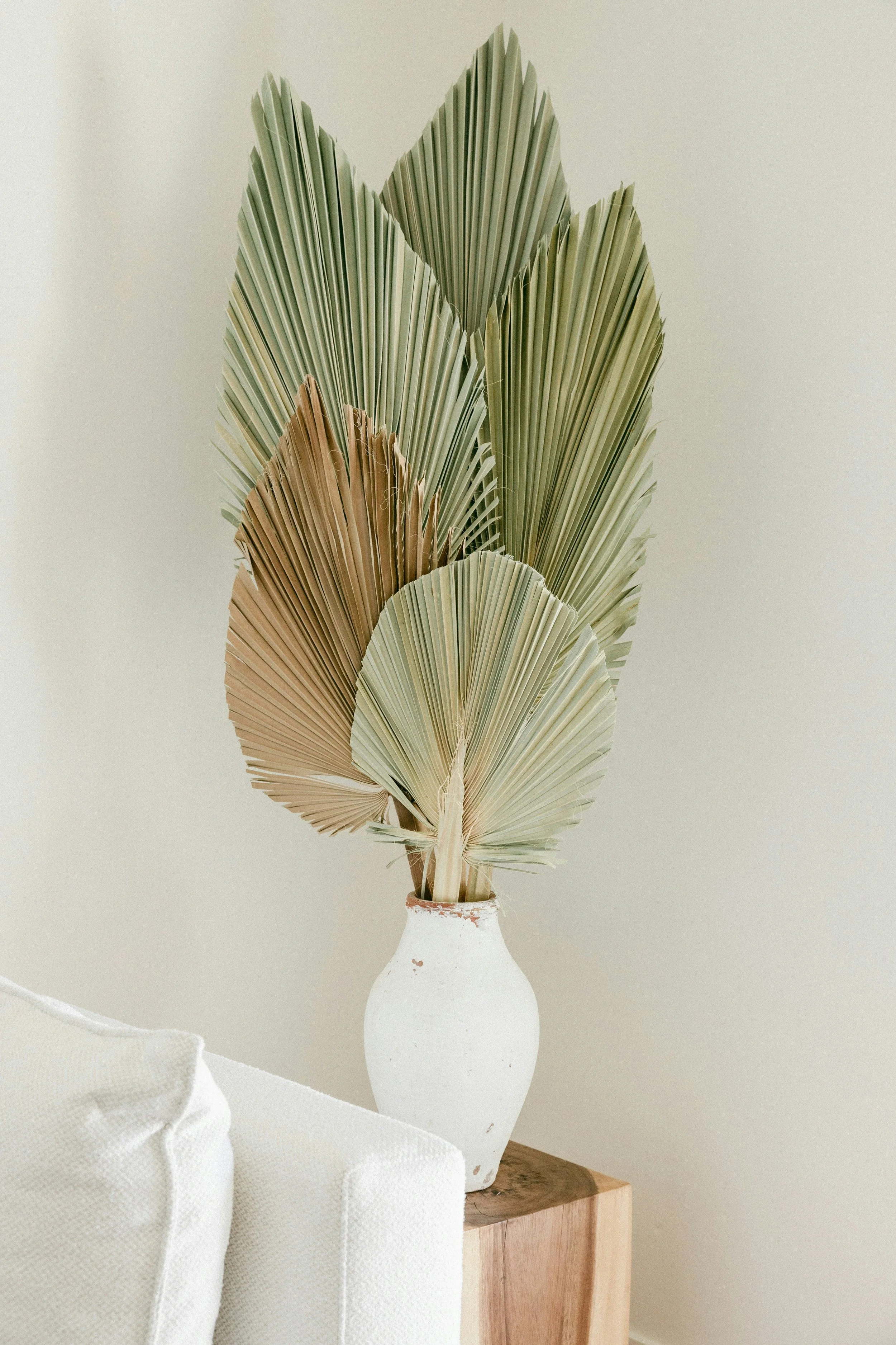 A white vase with a rustic finish holding dried palm leaves in shades of green, beige, and brown, placed on a wooden surface with part of a white upholstered piece of furniture visible in the foreground.