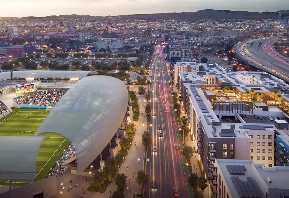 The Standard at Los Angeles – an aerial view of the city at dusk featuring a large stadium with a partially covered roof on the left, a busy road with moving vehicles, and modern buildings on the right side.