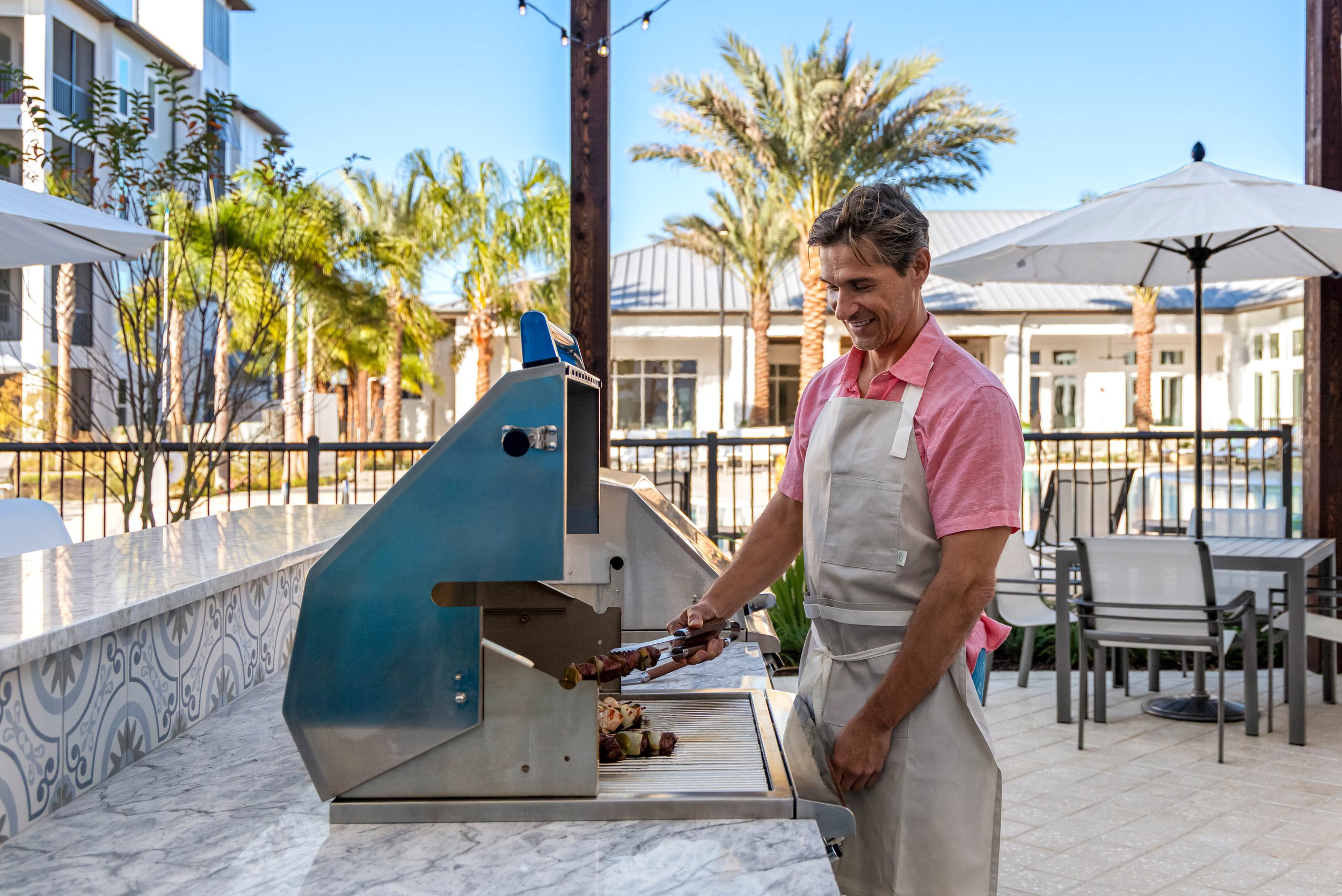 A man in a pink shirt and white apron grilling skewers outdoors at a restaurant with palm trees, umbrellas, and patio furniture in the background.