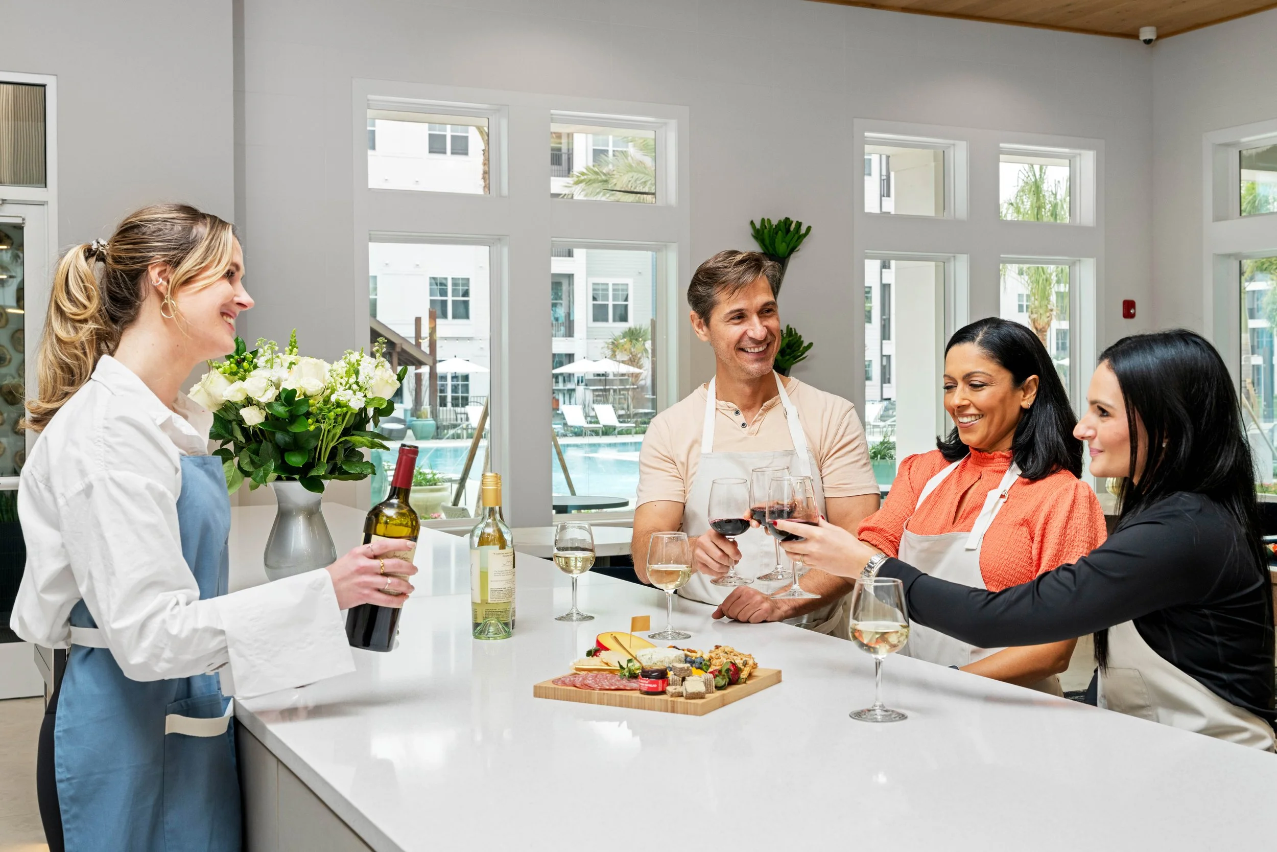 Four people enjoying a gathering in a bright kitchen with a view of a pool outside, sharing wine and celebrating next to a cheese and charcuterie board.