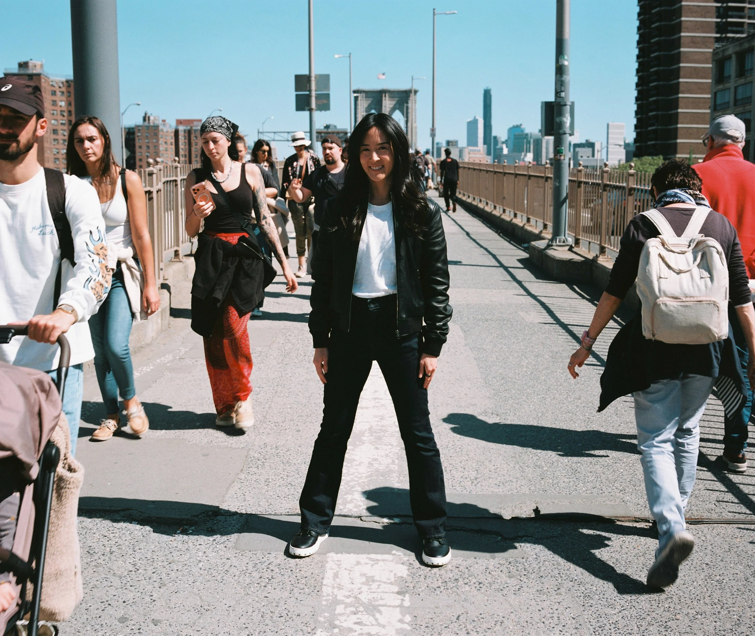 Maeve Wang standing on the Brooklyn Bridge in her IAMBIC shoes amidst crowds of people walking it