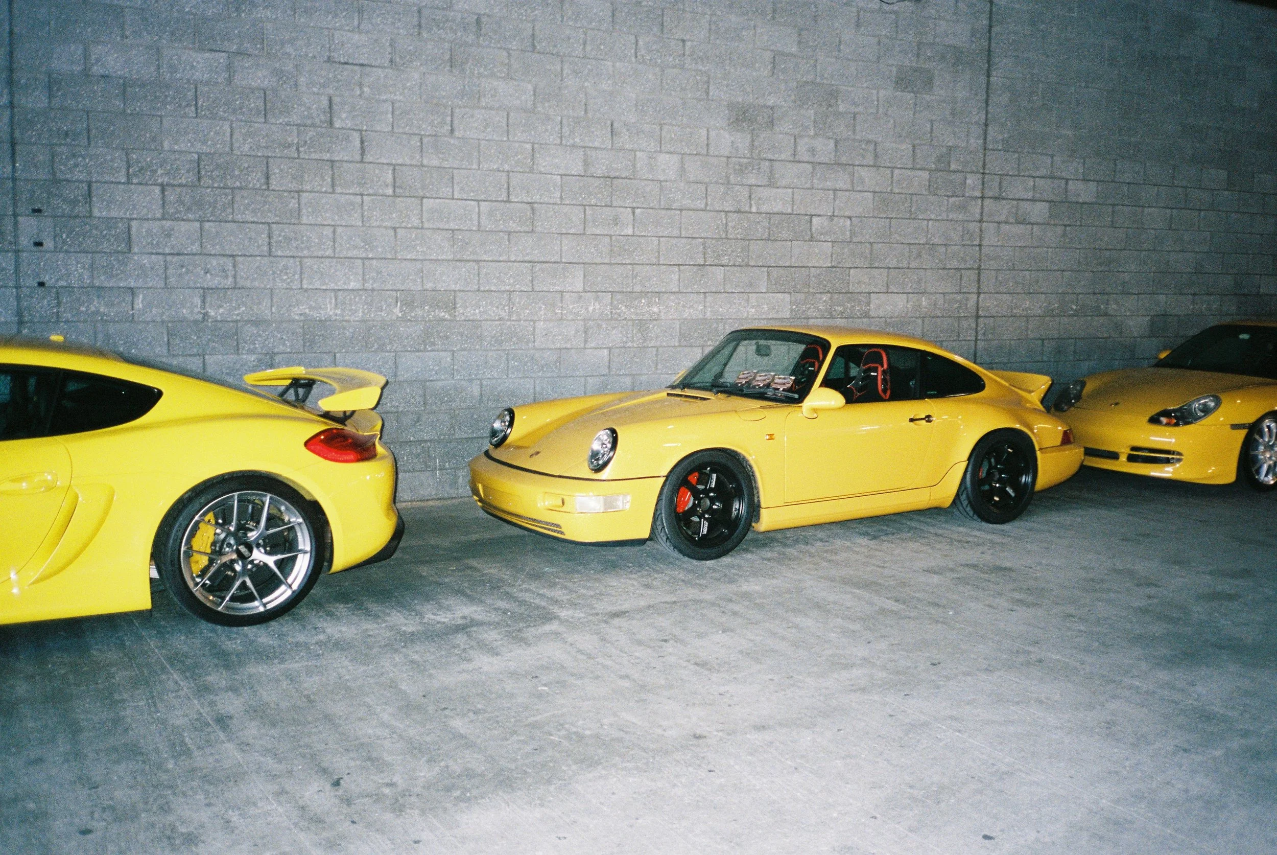 Film flash photography side view of three yellow Porsches in a line with a brick wall as backdrop