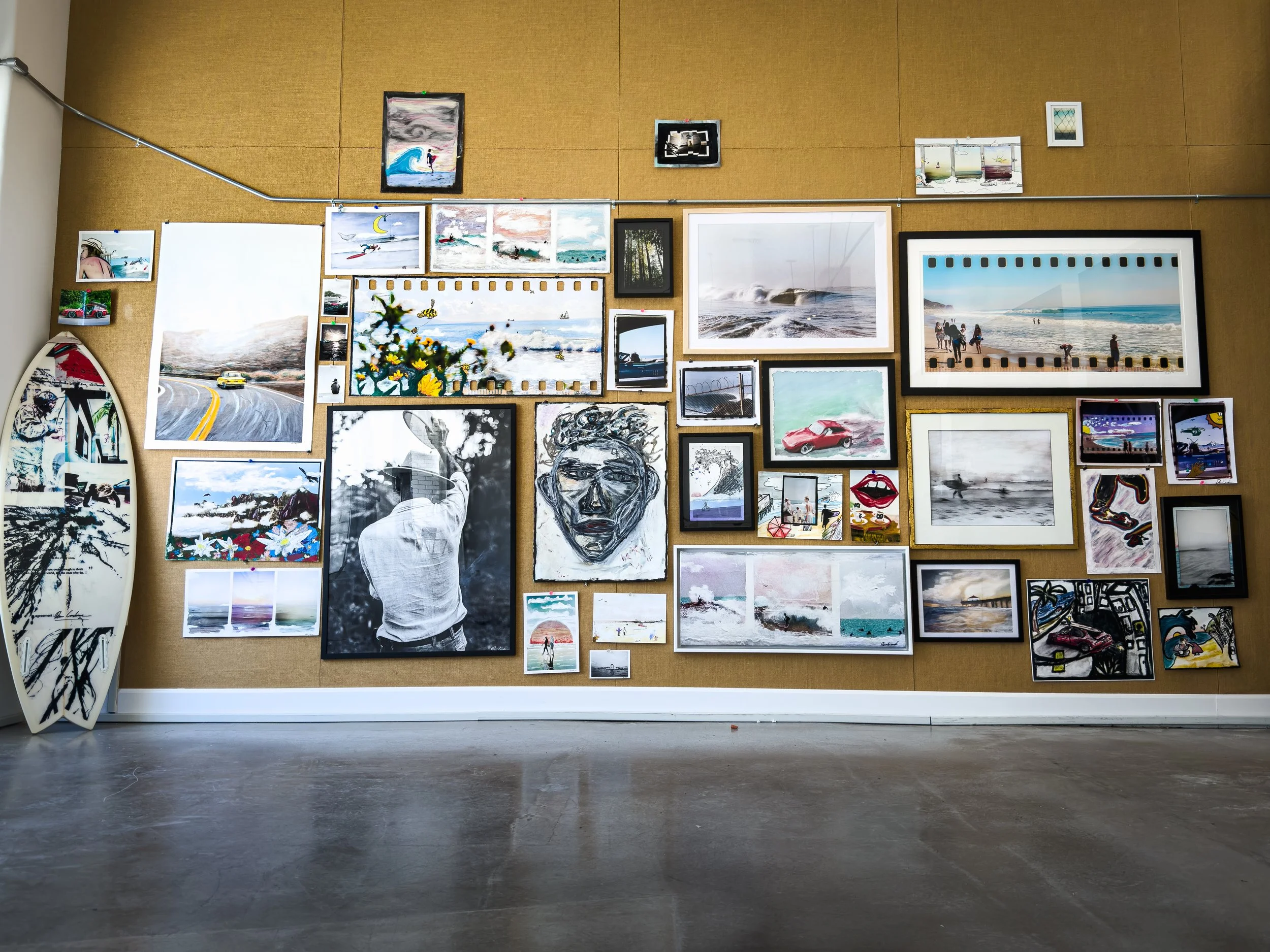 A wall at a coffee shop popup art show displaying the author's photography and mixed media work in various framed prints and one piece adorning a surfboard leaning up against the wall