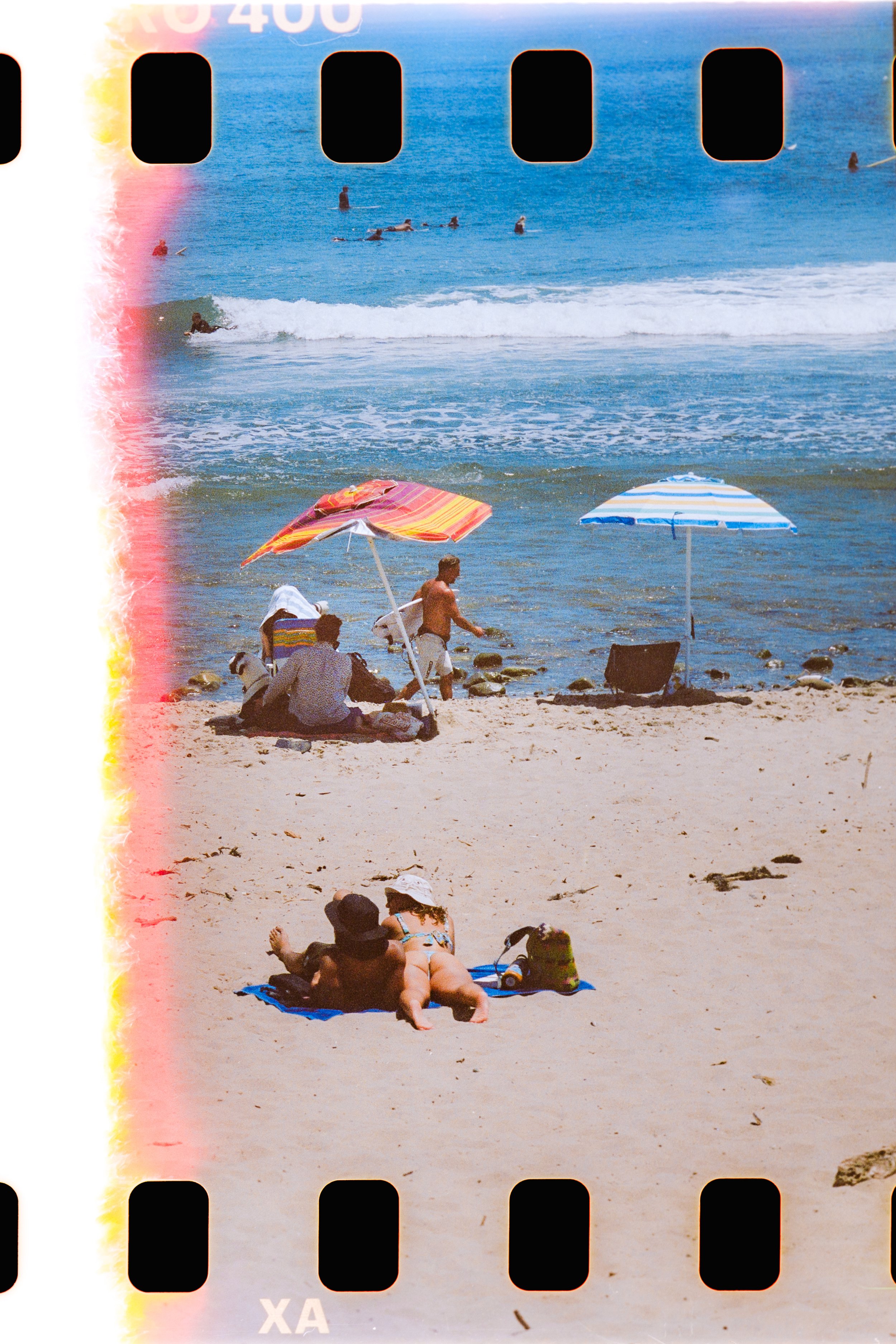 A photographic of sunbathers on a beach looking out onto surfers waiting for waves and surfing. The lefthand side of the frame shows a light leak