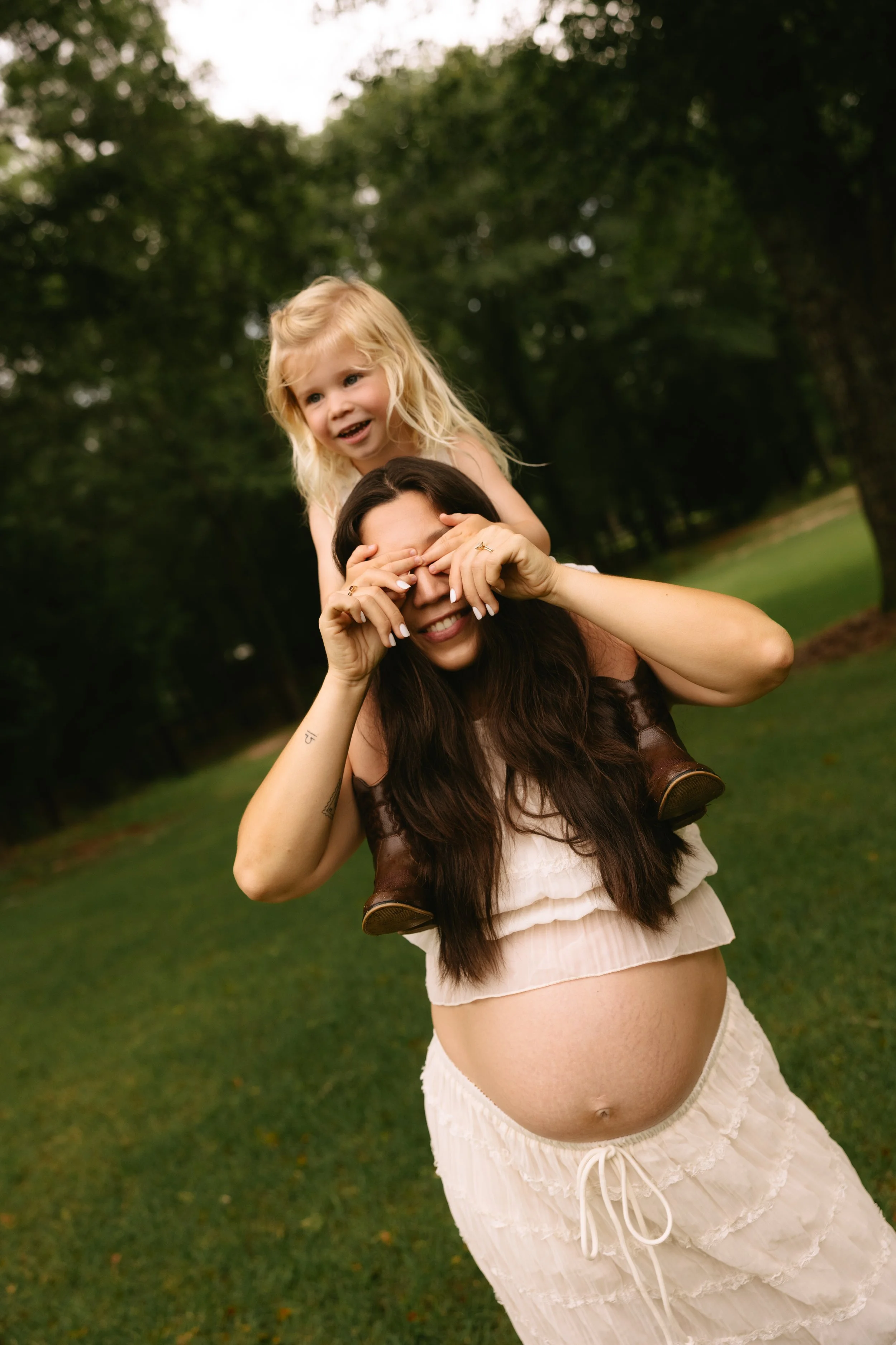 A pregnant woman with long dark hair giving a children piggyback ride in a park, with a young girl with blond hair sitting on her shoulders, both smiling and enjoying a sunny day.