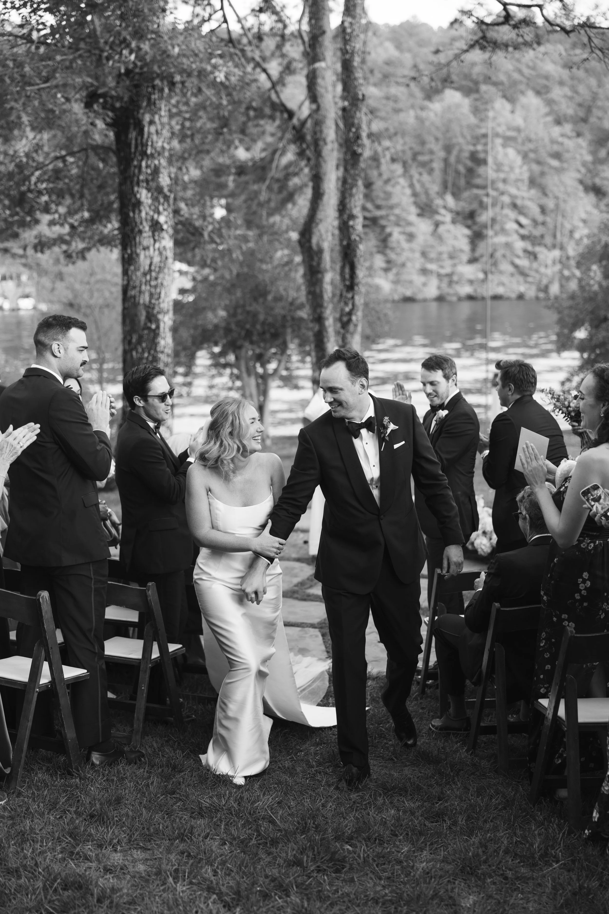 A black and white photo of a wedding ceremony, with a groom and bride walking hand in hand down the aisle, surrounded by guests clapping and smiling. The outdoor setting features trees and a lake in the background.