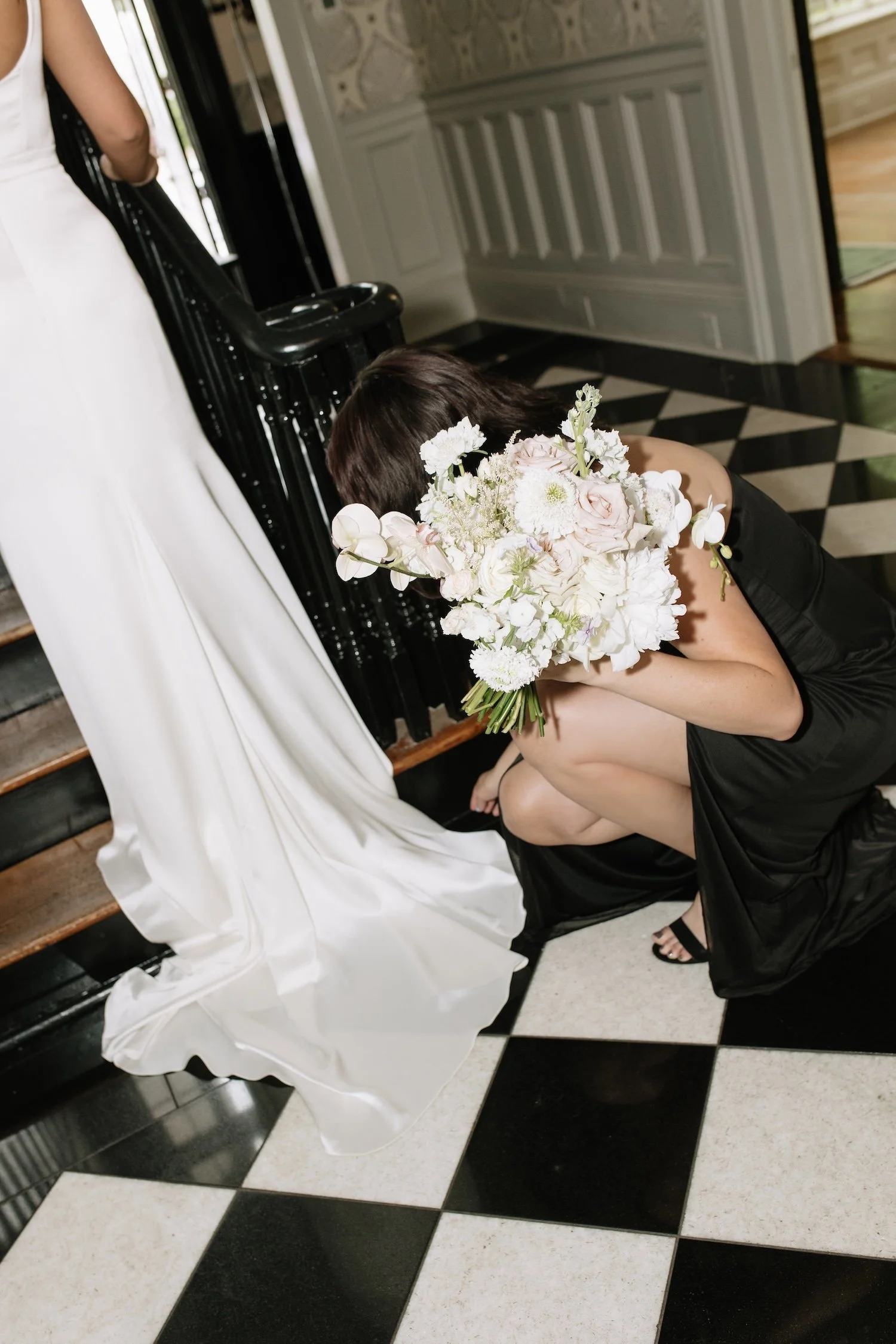 A woman with shoulder-length dark hair, dressed in a black sleeveless dress, crouching on a black-and-white checkered floor, holding a large bouquet of white and pale pink flowers while partially hidden behind it. The scene appears to be indoors, nea