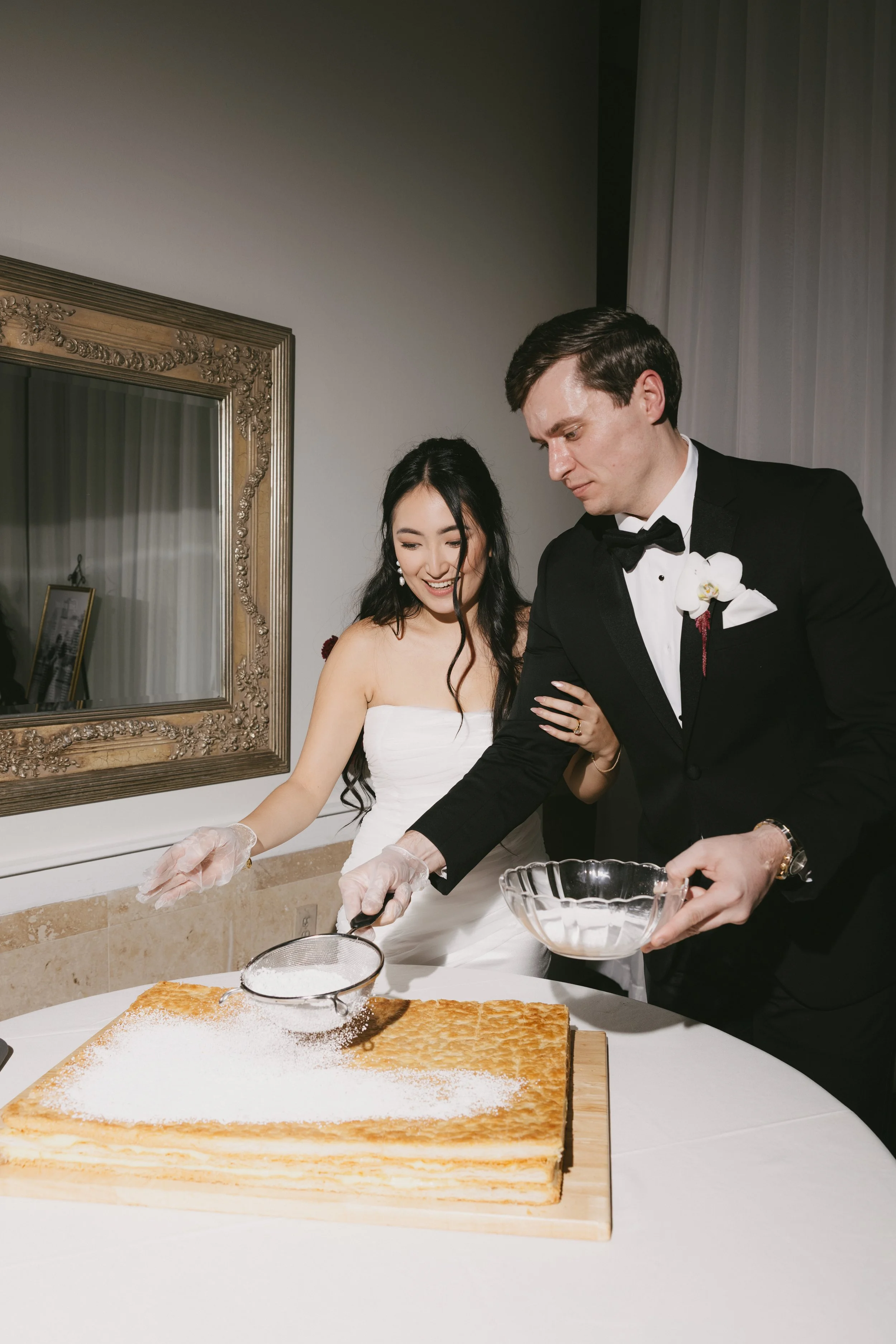 A bride and groom cutting a tiered wedding cake together, wearing gloves, with the bride smiling and the groom focused on cutting.