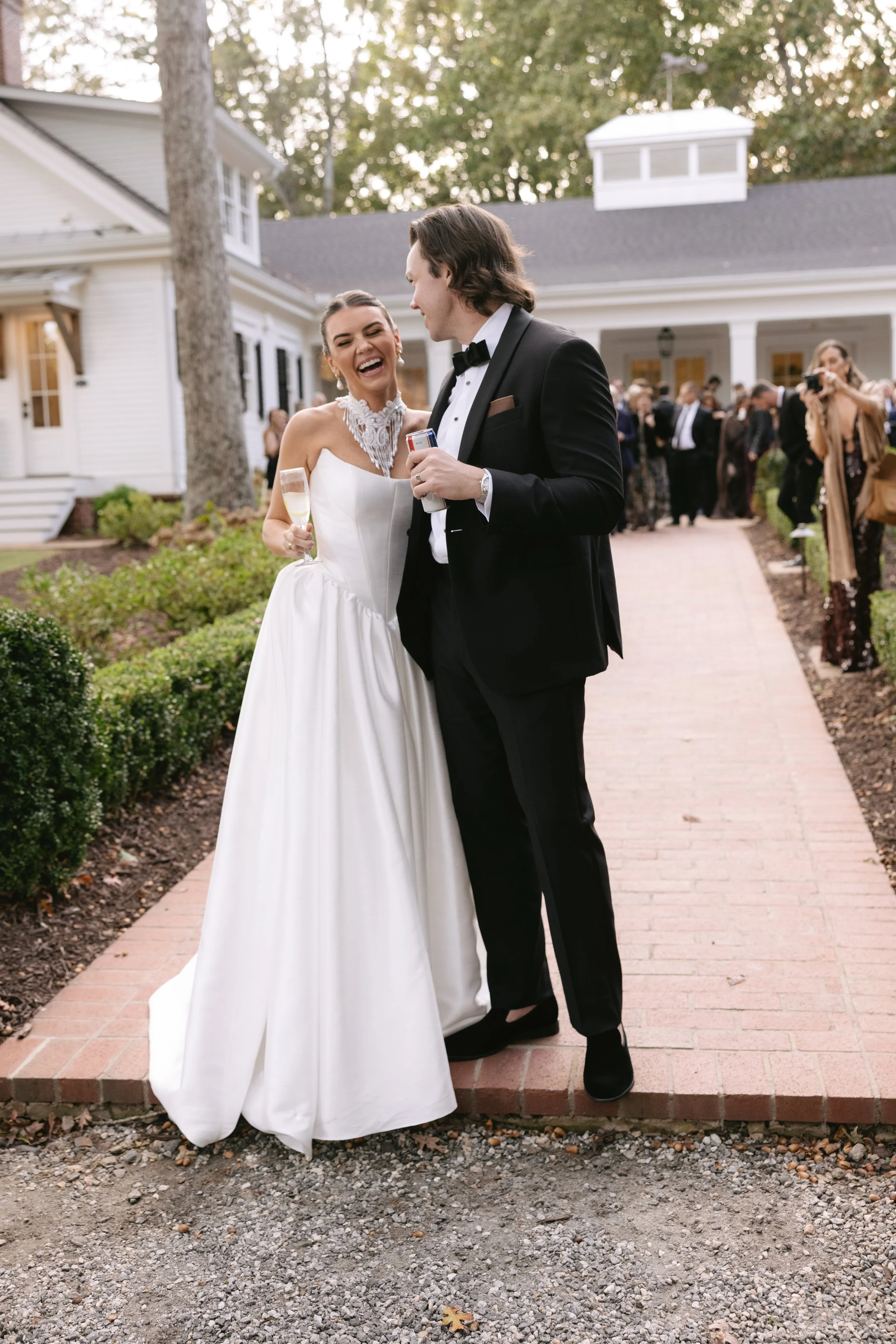 A bride and groom laughing and sharing a moment during their wedding celebration outside. The bride is in a white wedding dress and the groom is in a black tuxedo. They are surrounded by guests and the scene takes place on a brick pathway near a whit