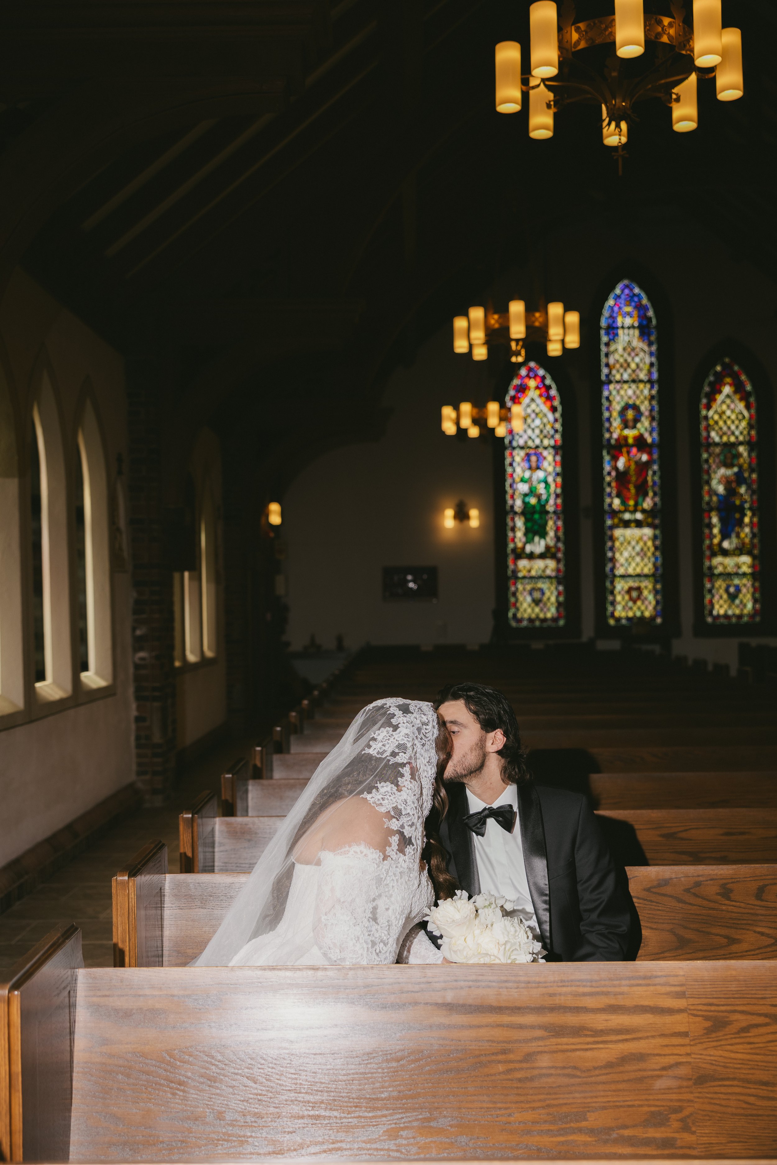 A bride and groom share a kiss in a church, with stained glass windows and hanging lights visible in the background.