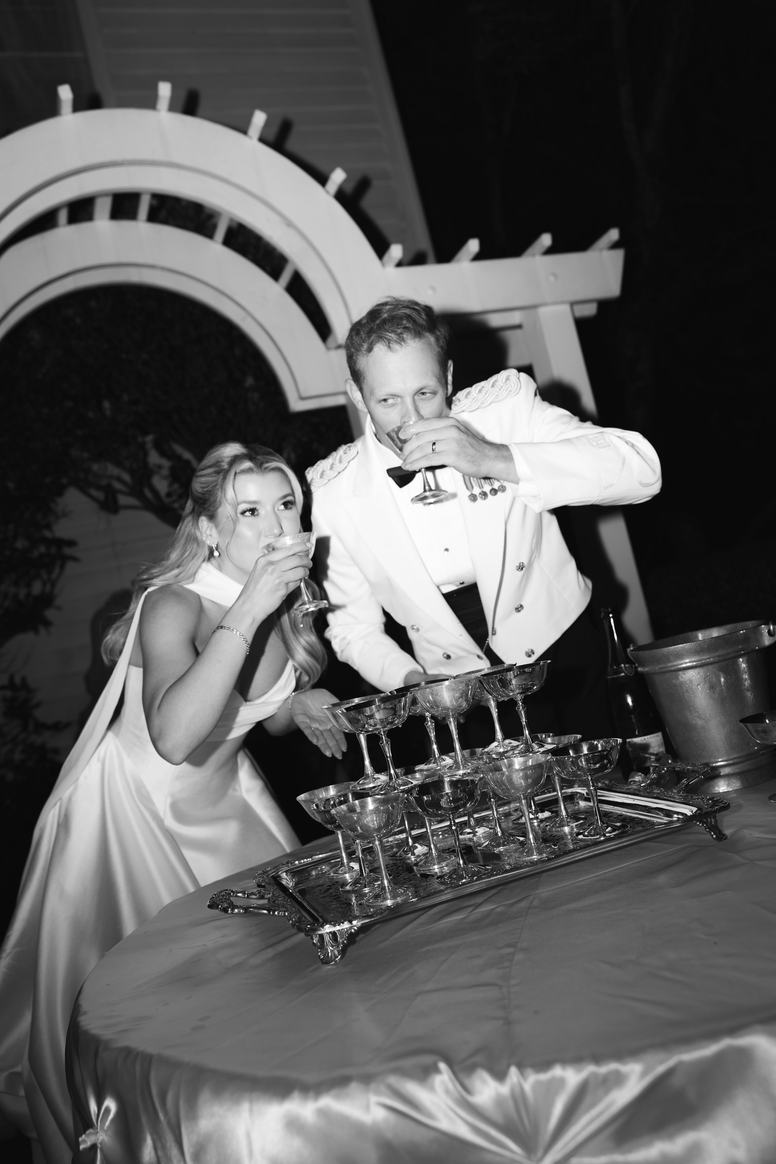 A woman and a man in formal attire, pouring drinks into champagne glasses on a table.