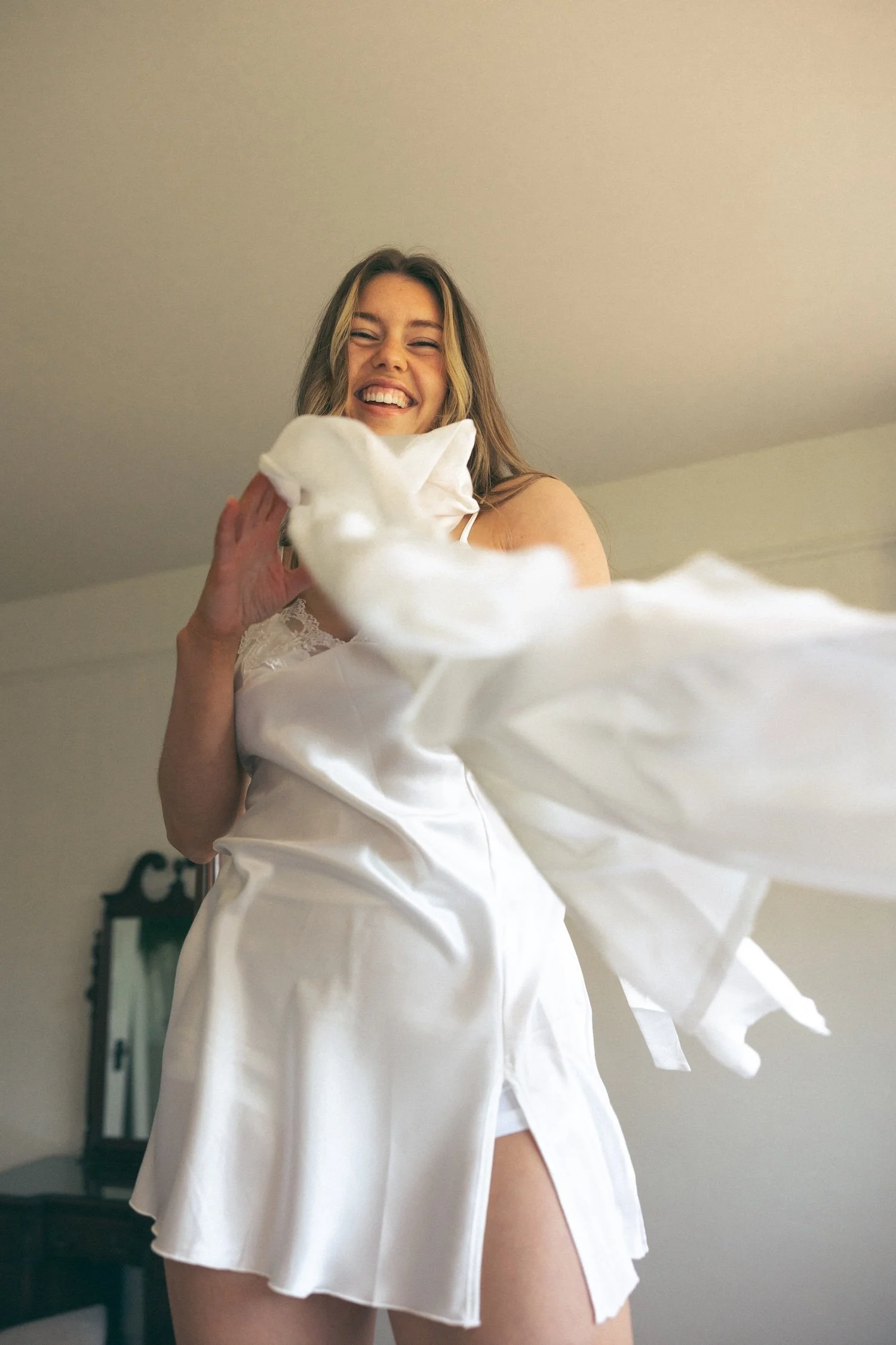 A woman in a white satin robe smiling joyfully as she playfully throws a pillow in a room with a mirror and neutral-colored walls.