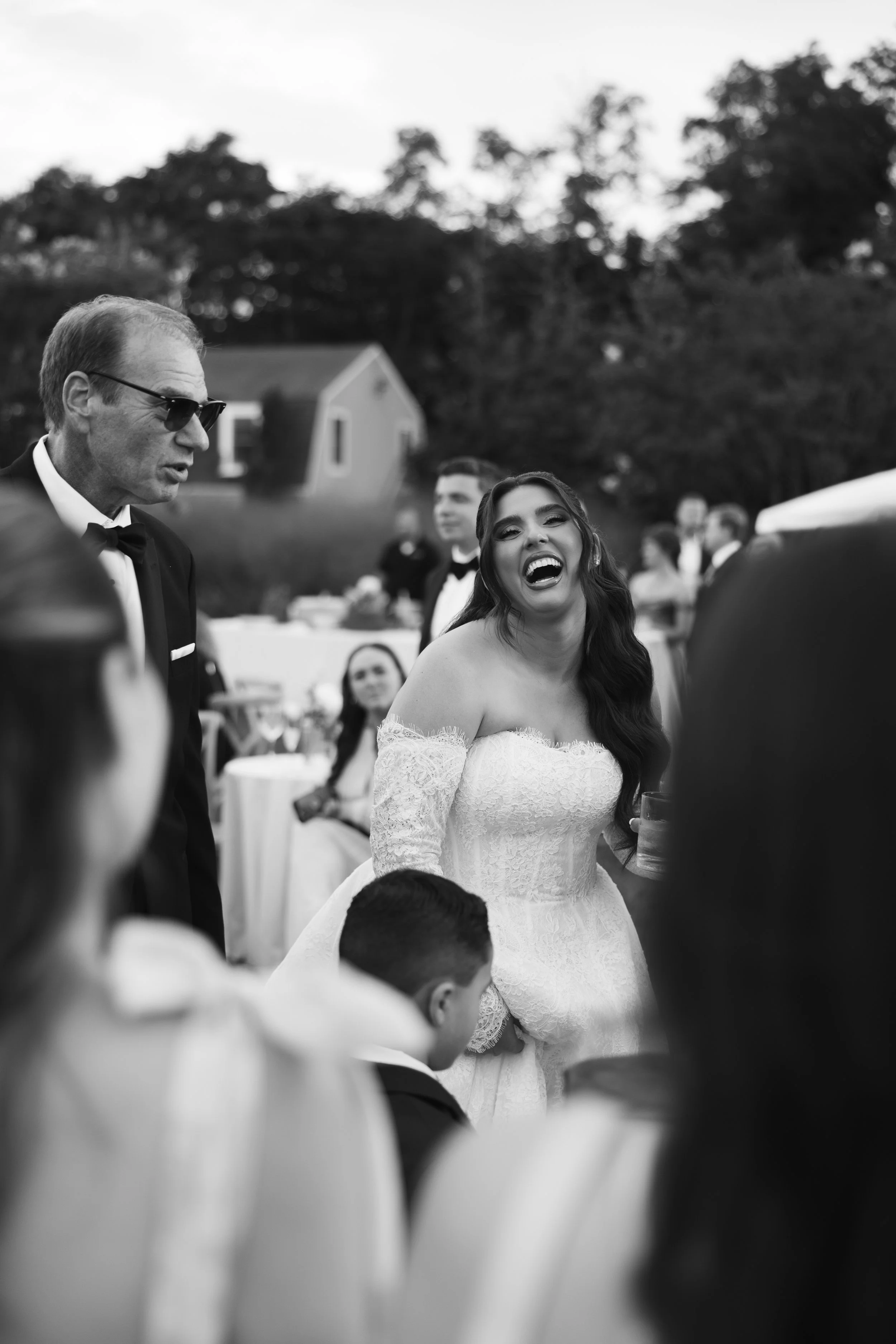 A black-and-white photo of a joyful bride laughing during a wedding reception outdoors, surrounded by guests including a man in sunglasses and a tuxedo, with trees and a house in the background.