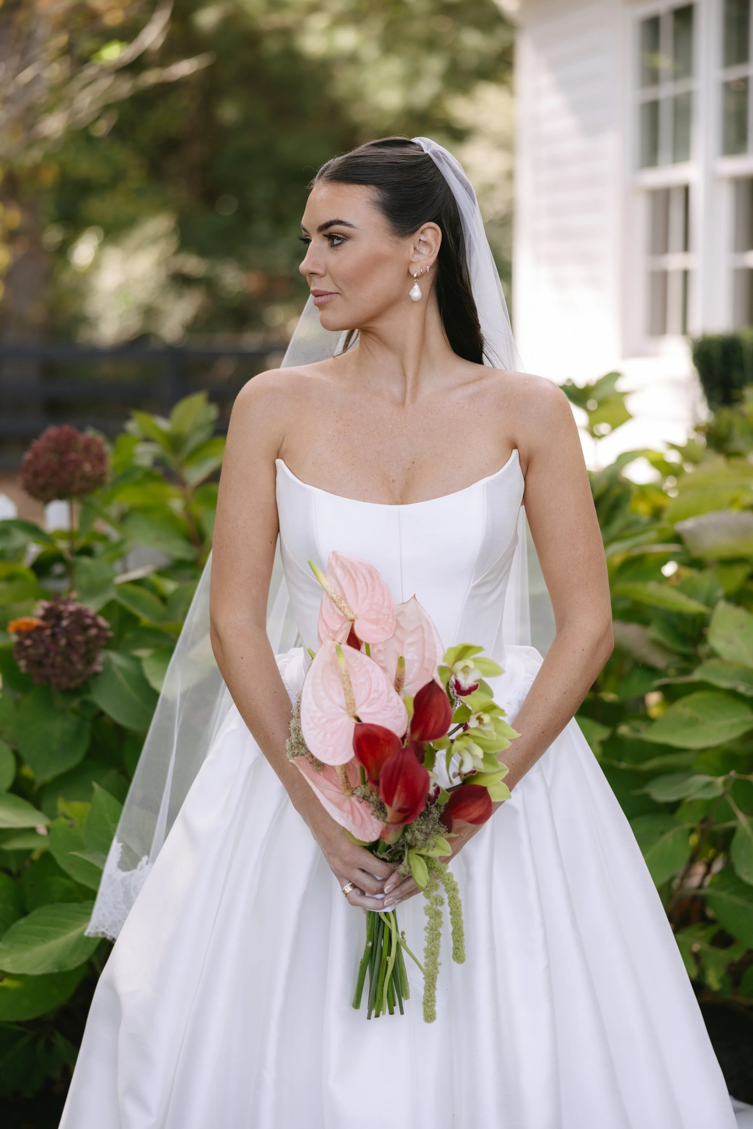A bride in a white wedding gown holds a bouquet of pink and red flowers, standing outdoors in a garden setting.