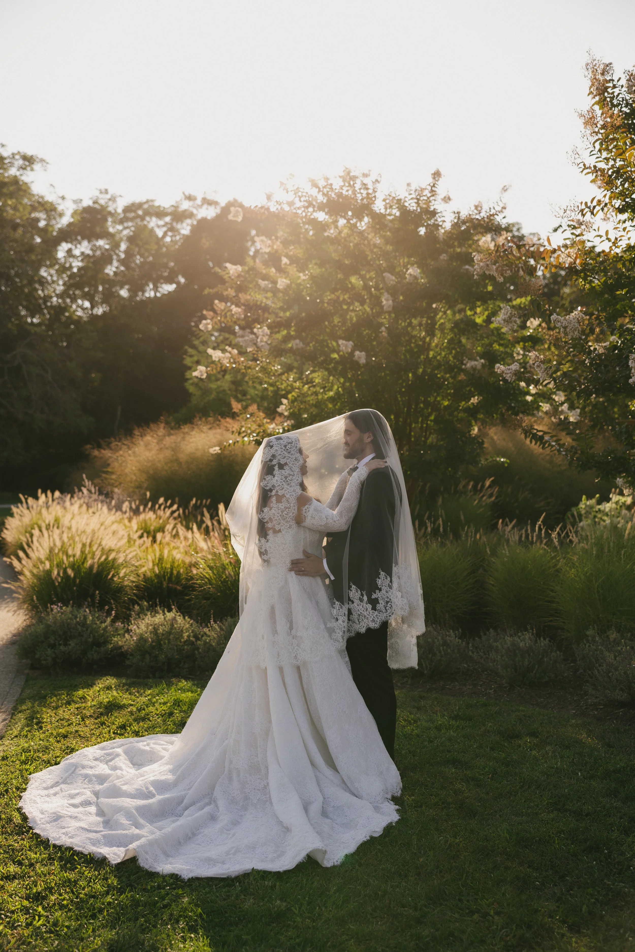 A bride and groom in wedding attire standing outdoors, facing each other under a sheer veil, surrounded by greenery and flowers, with sunlight creating a warm glow.