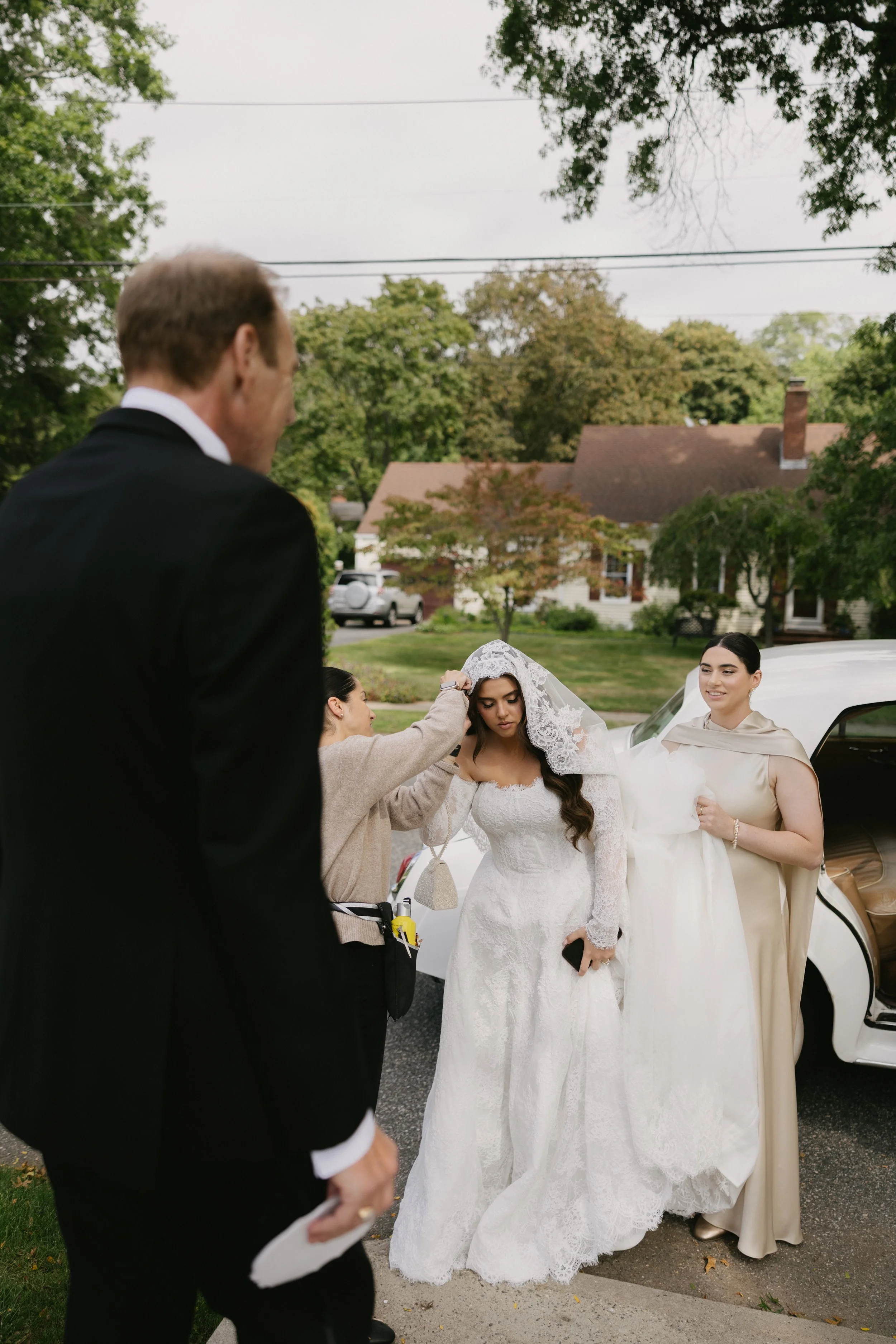 A bride in a white lace wedding gown getting ready with assistance from her friends or family outside a house with trees in the background. A limousine is parked nearby.