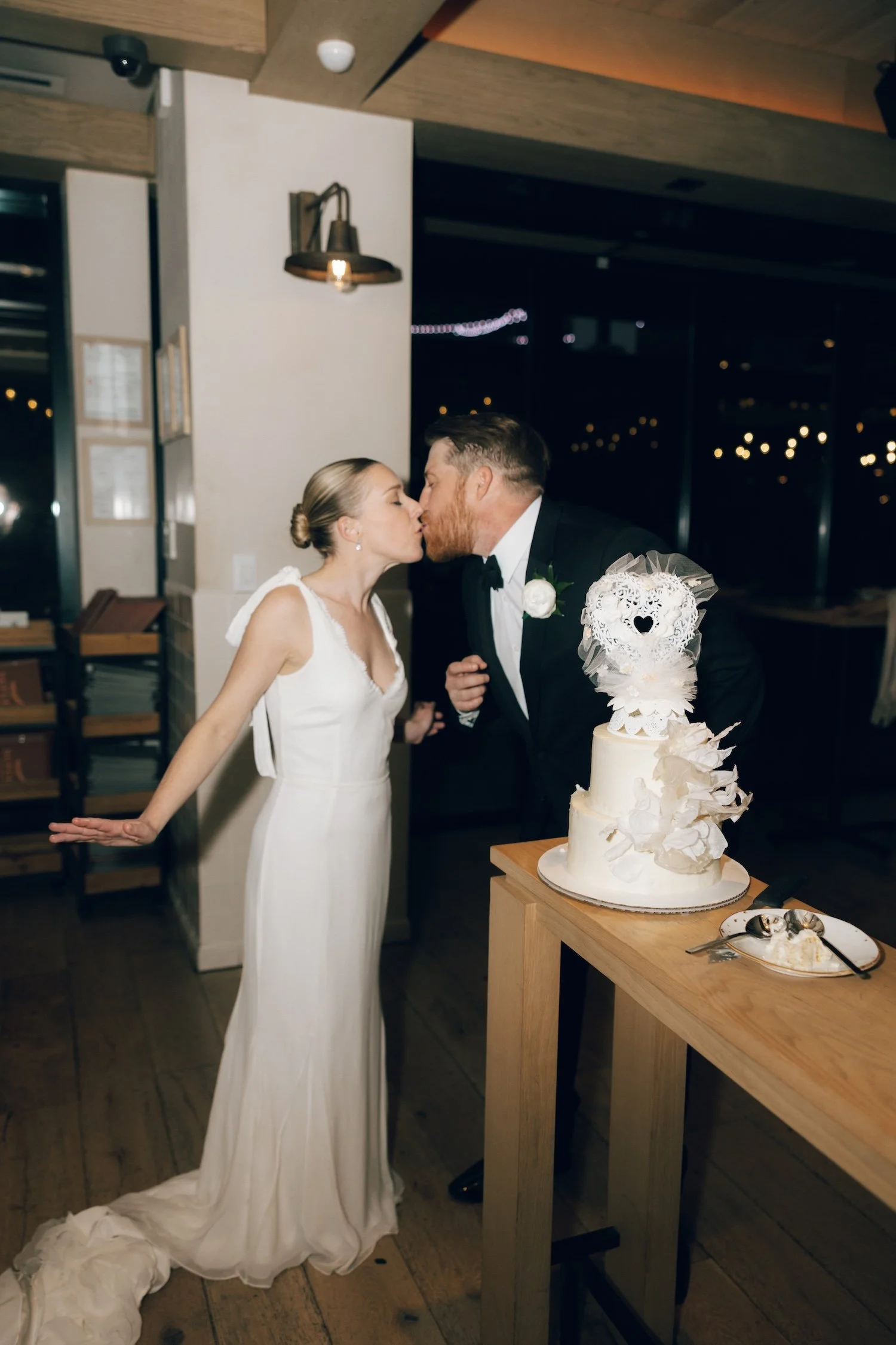 Bride and groom kissing at their wedding reception, with a wedding cake on a table nearby.