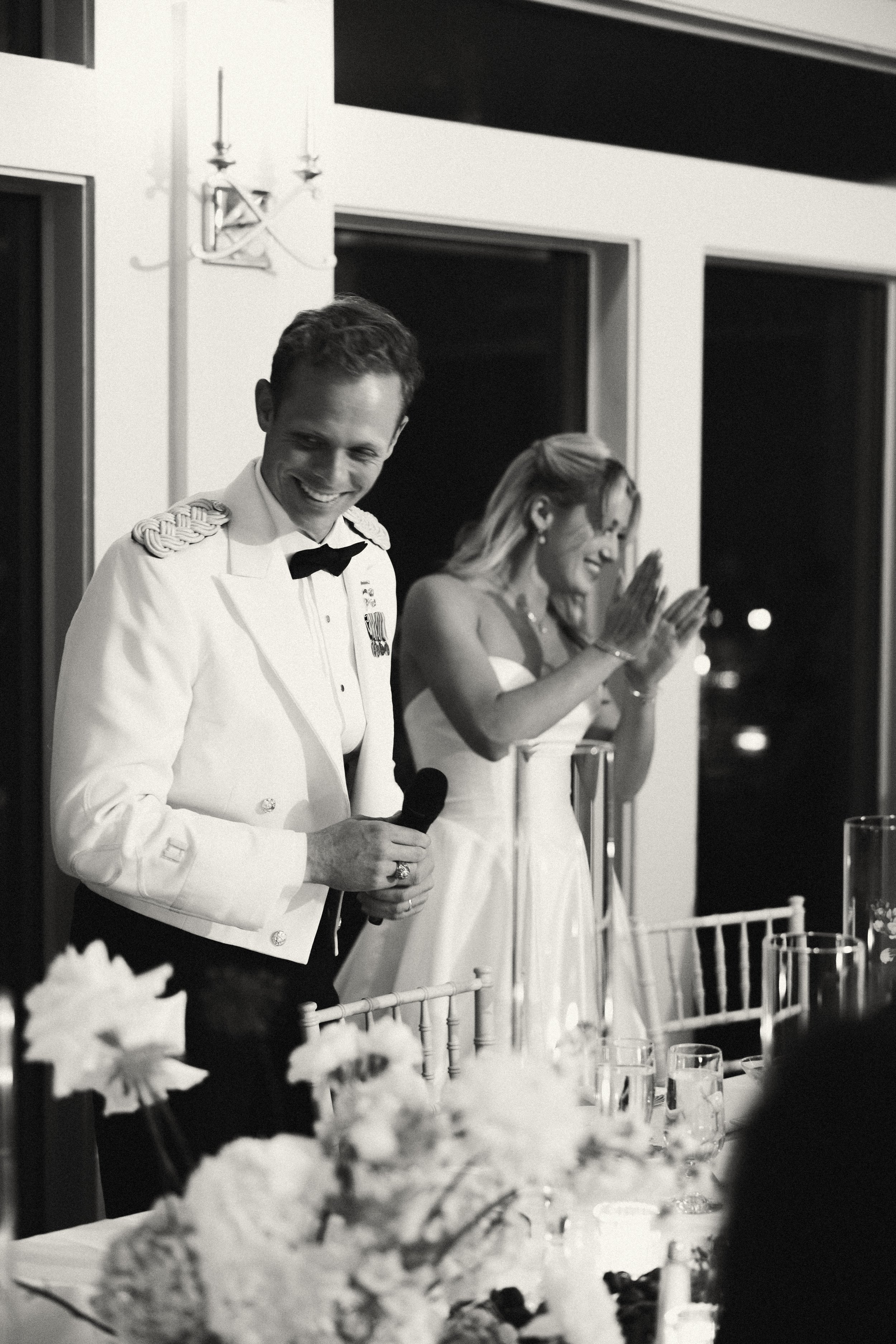 A black-and-white photo of a man in a military uniform holding a microphone, standing next to a woman clapping during a celebration or speech at an indoor event.