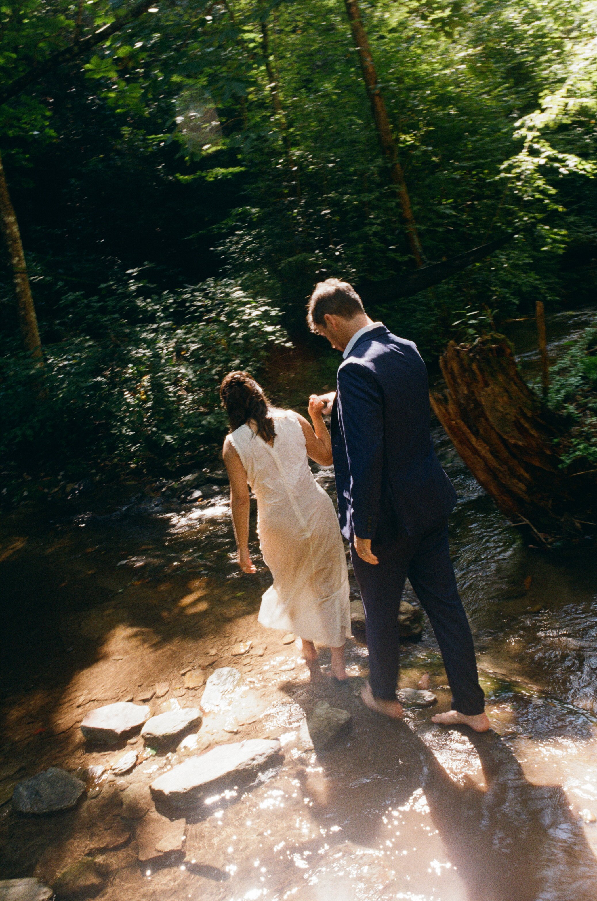 A couple, with the woman in a white dress and the man in a dark suit, walking barefoot through a shallow creek in a lush forest.