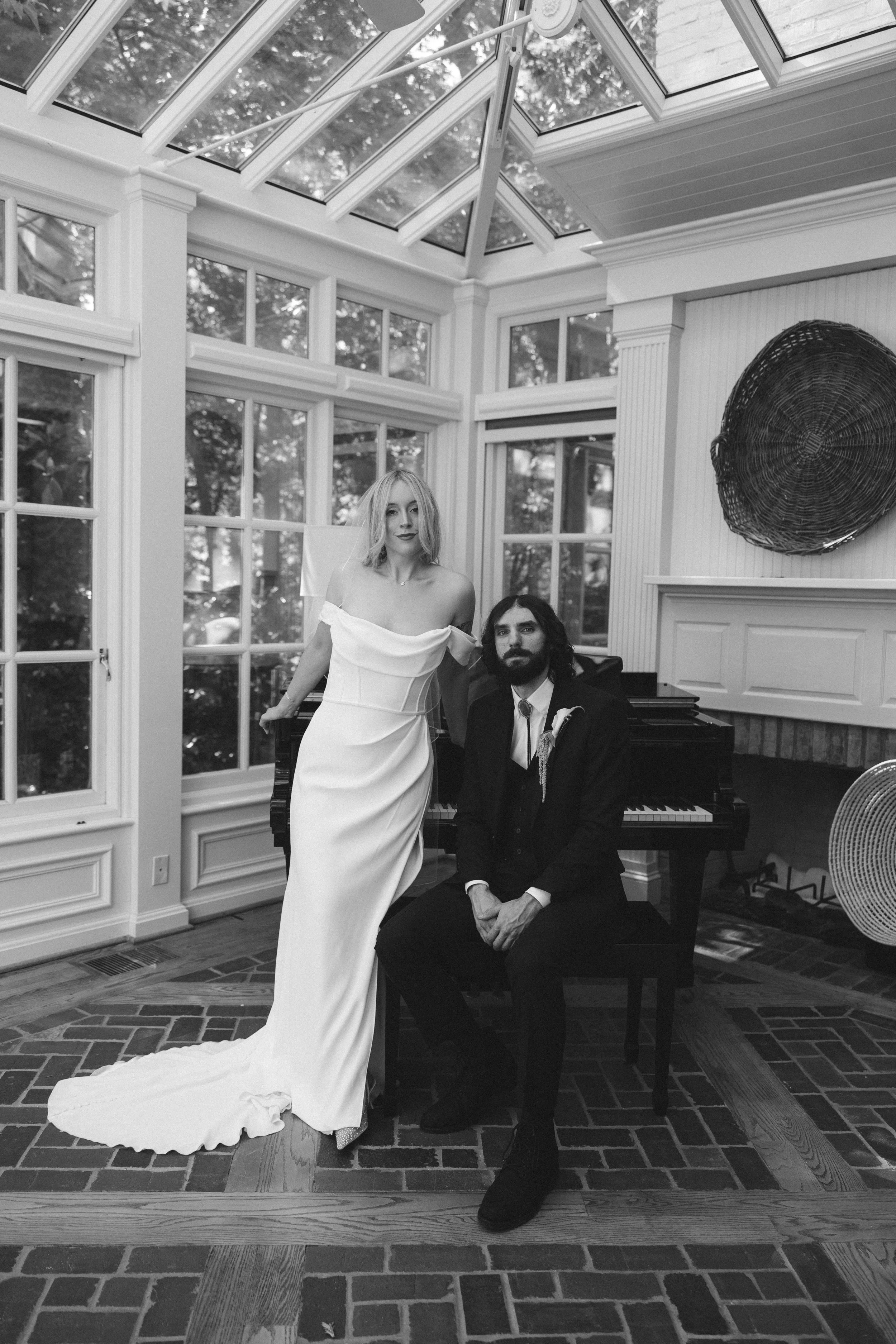 A man in formal attire sitting at a piano and a woman in a white wedding dress standing beside him in a bright room with large windows and a glass ceiling.