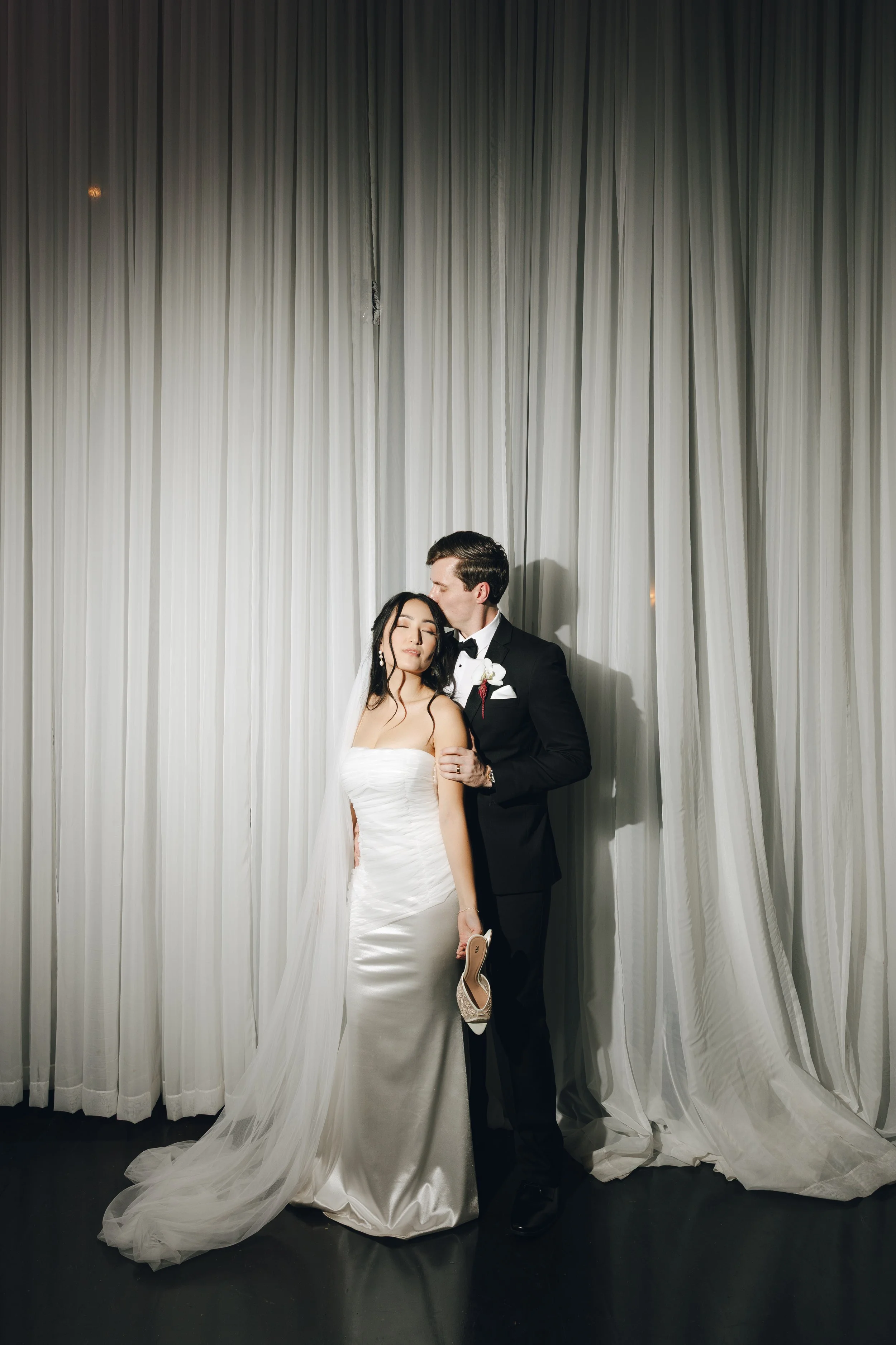 Bride and groom on wedding day, standing in front of white curtains, with the groom gently kissing the bride's forehead.