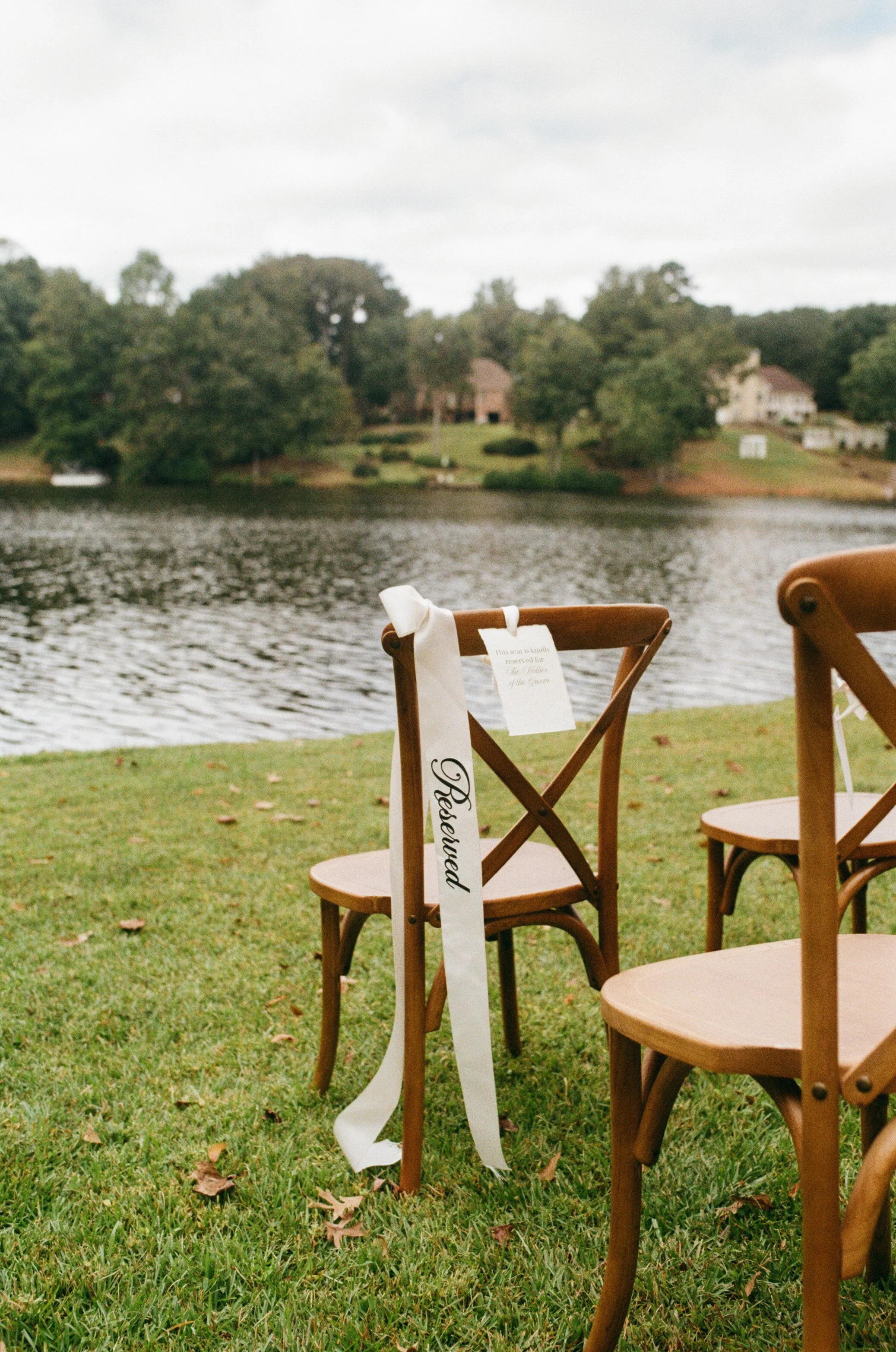 Wooden chairs arranged outdoors by a lake, with a 'Reserved' ribbon on one chair and a white tag, suggesting a wedding or special event setup.