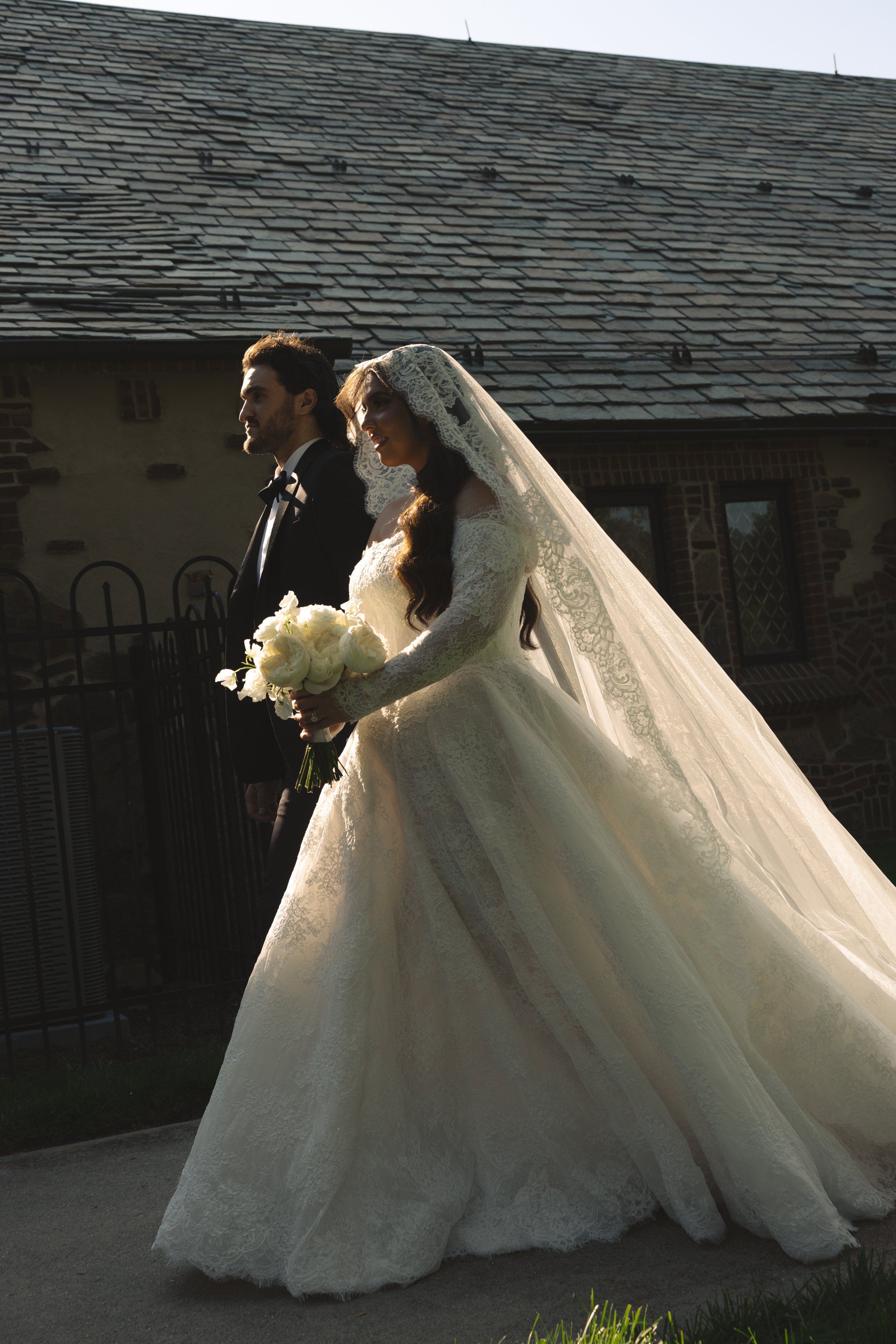 Bride and groom walking outside, with the bride in a long, lace wedding gown holding a bouquet of white roses, and the groom in a black tuxedo, backlit by the sun.