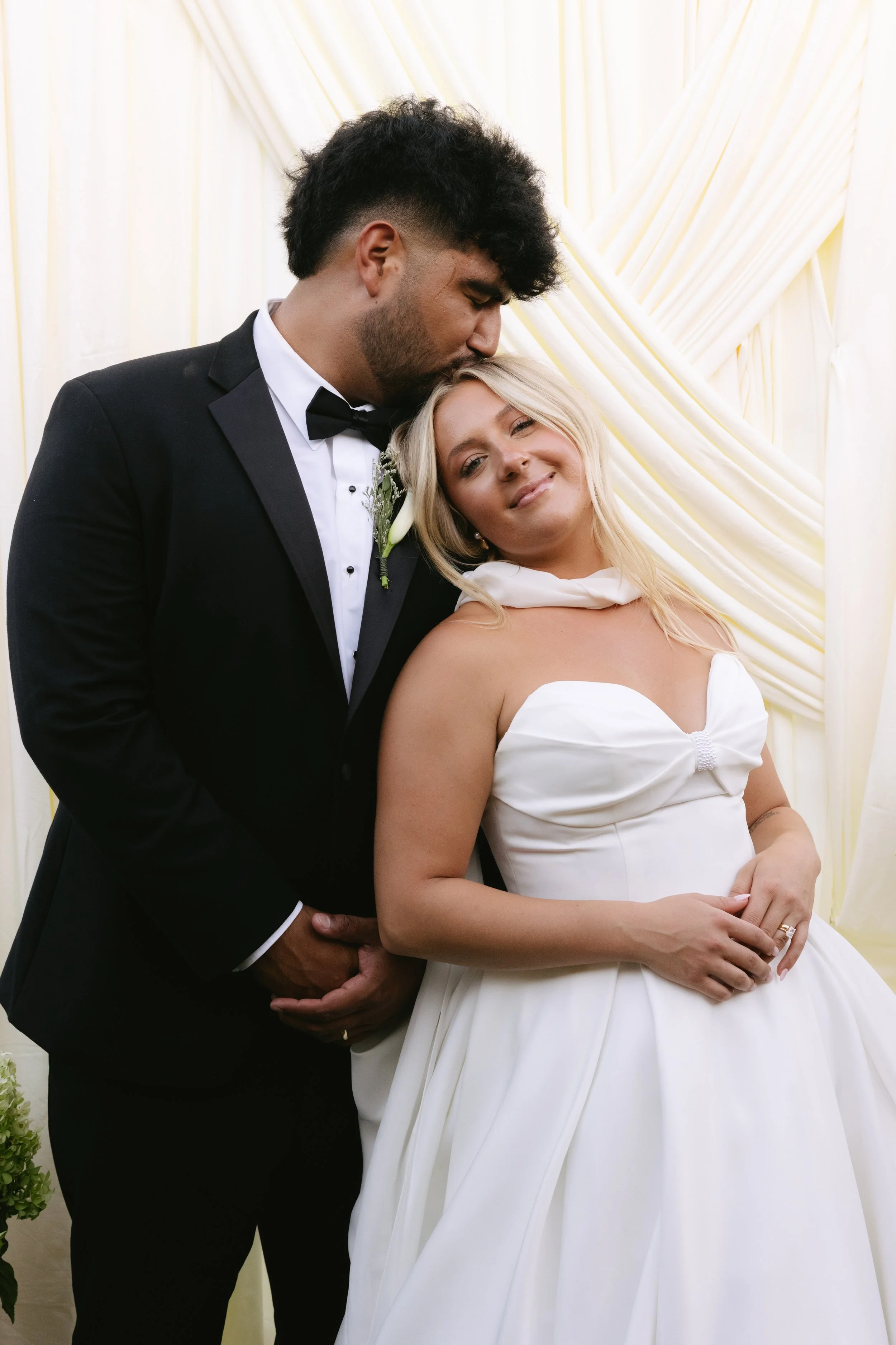 A groom in a black tuxedo and bow tie kissing a smiling bride in a white gown on her head and shoulder, standing in front of cream-colored drapes.
