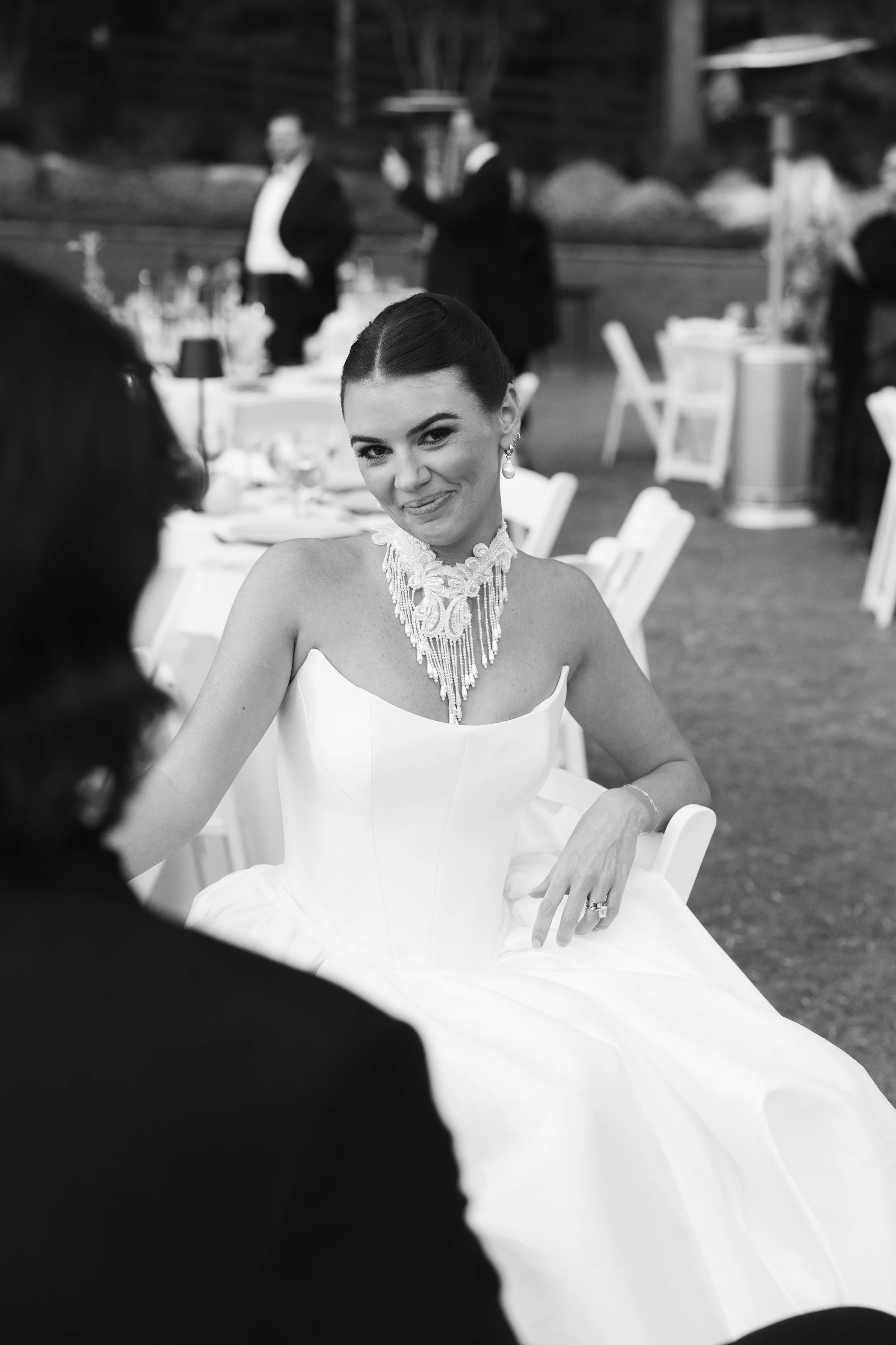 A woman in a wedding dress smiling at a wedding reception, with guests and tables visible in the background.