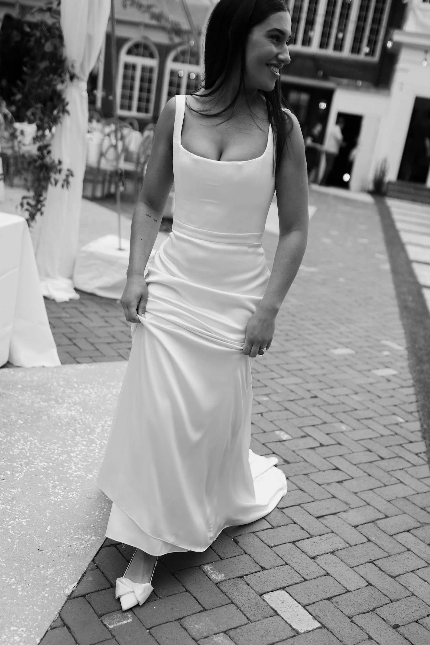 A woman in a sleeveless wedding dress adjusting her gown on a brick walkway at an indoor event.