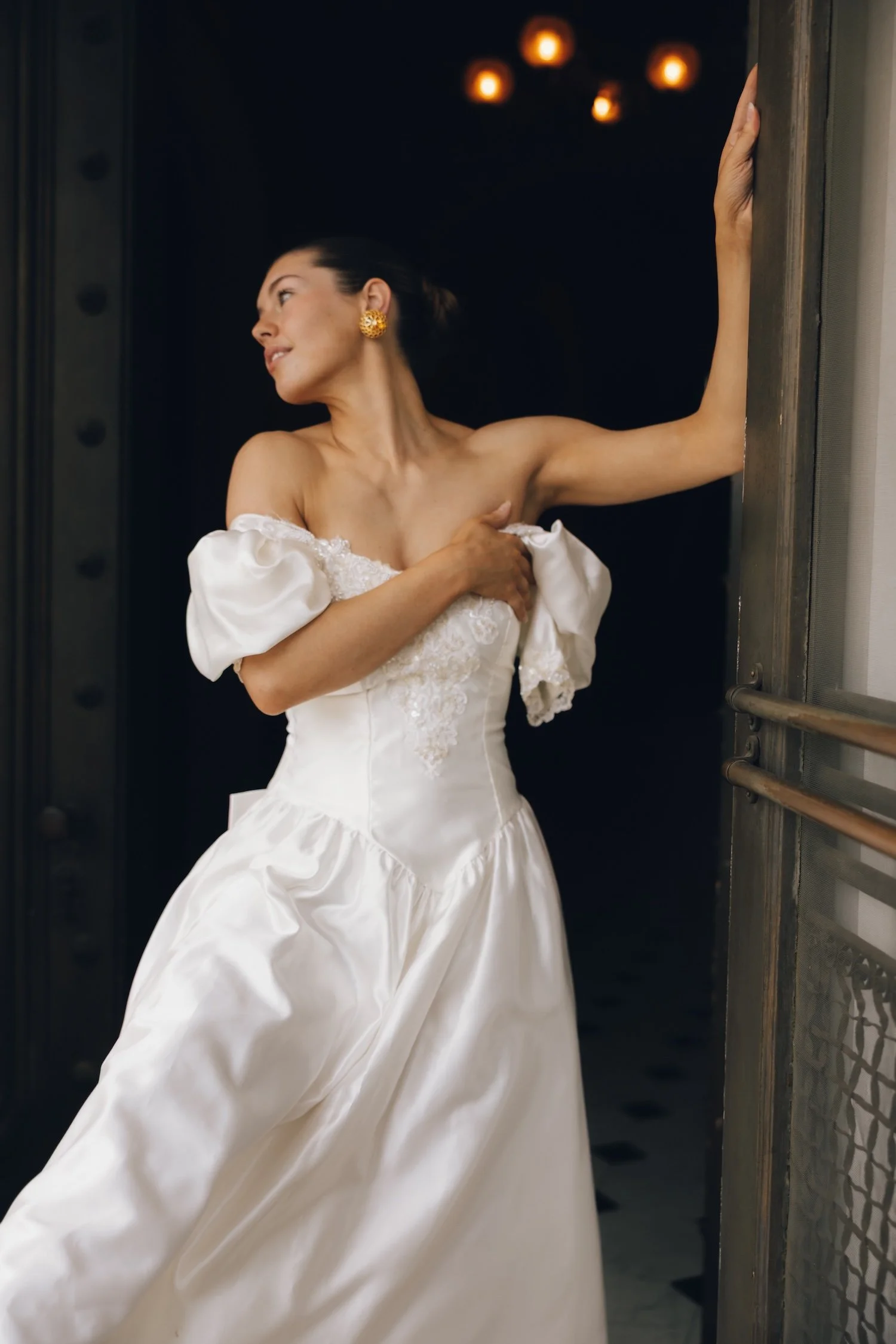 Woman in a white wedding dress with puffed sleeves, standing on a dance floor with her arm on a doorframe, looking away.