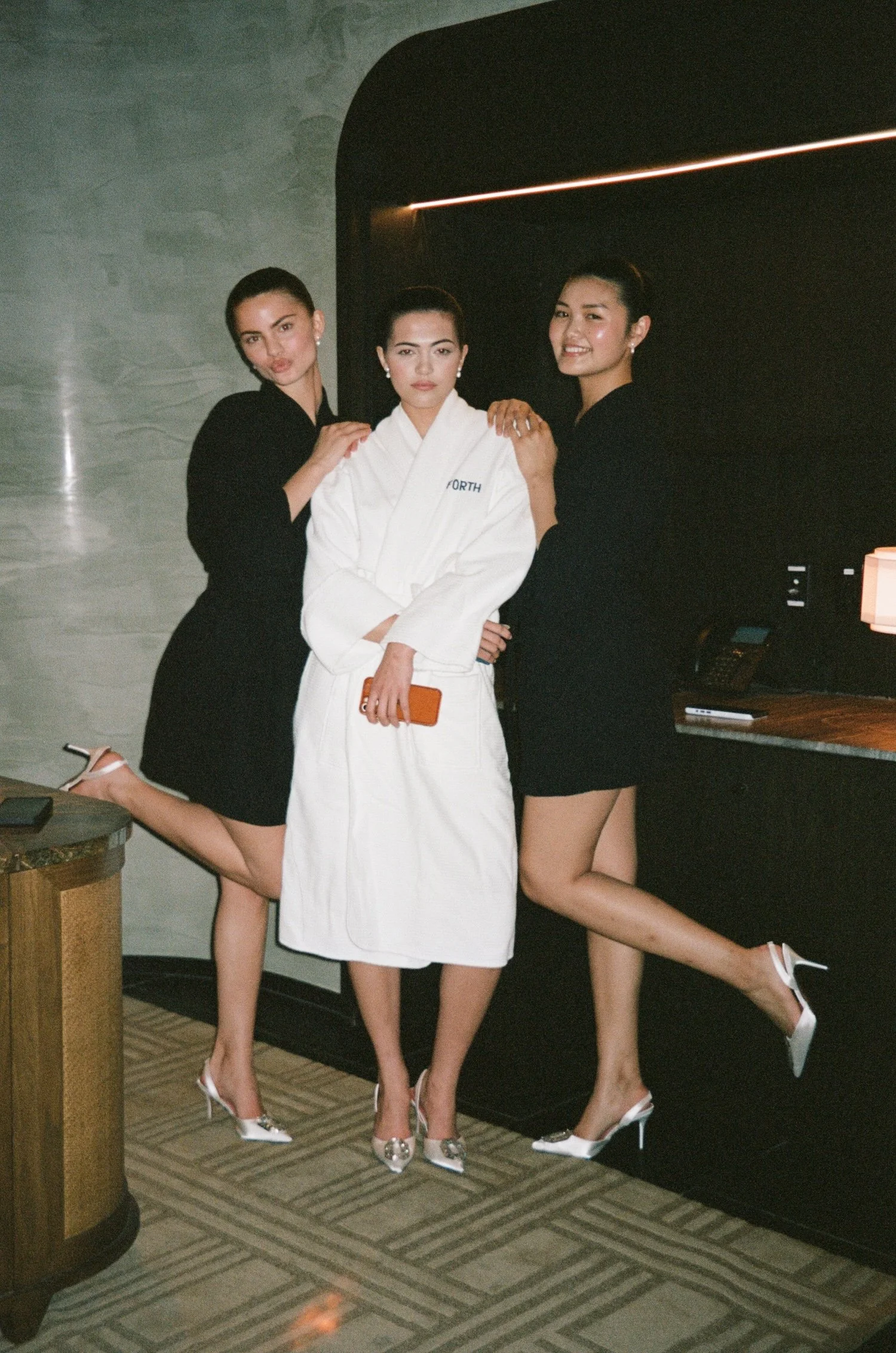 Three women posing indoors, two wearing black dresses and high heels, and one in a white bathrobe holding a phone.