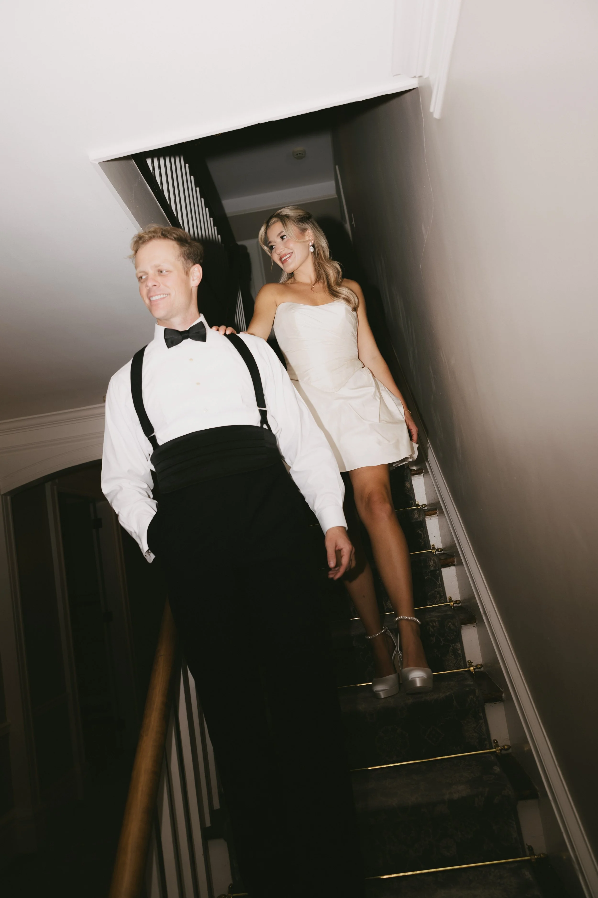 A couple dressed in formal attire walking down a staircase. The man is wearing a tuxedo with a bow tie, and the woman is wearing a wedding dress with high heels.