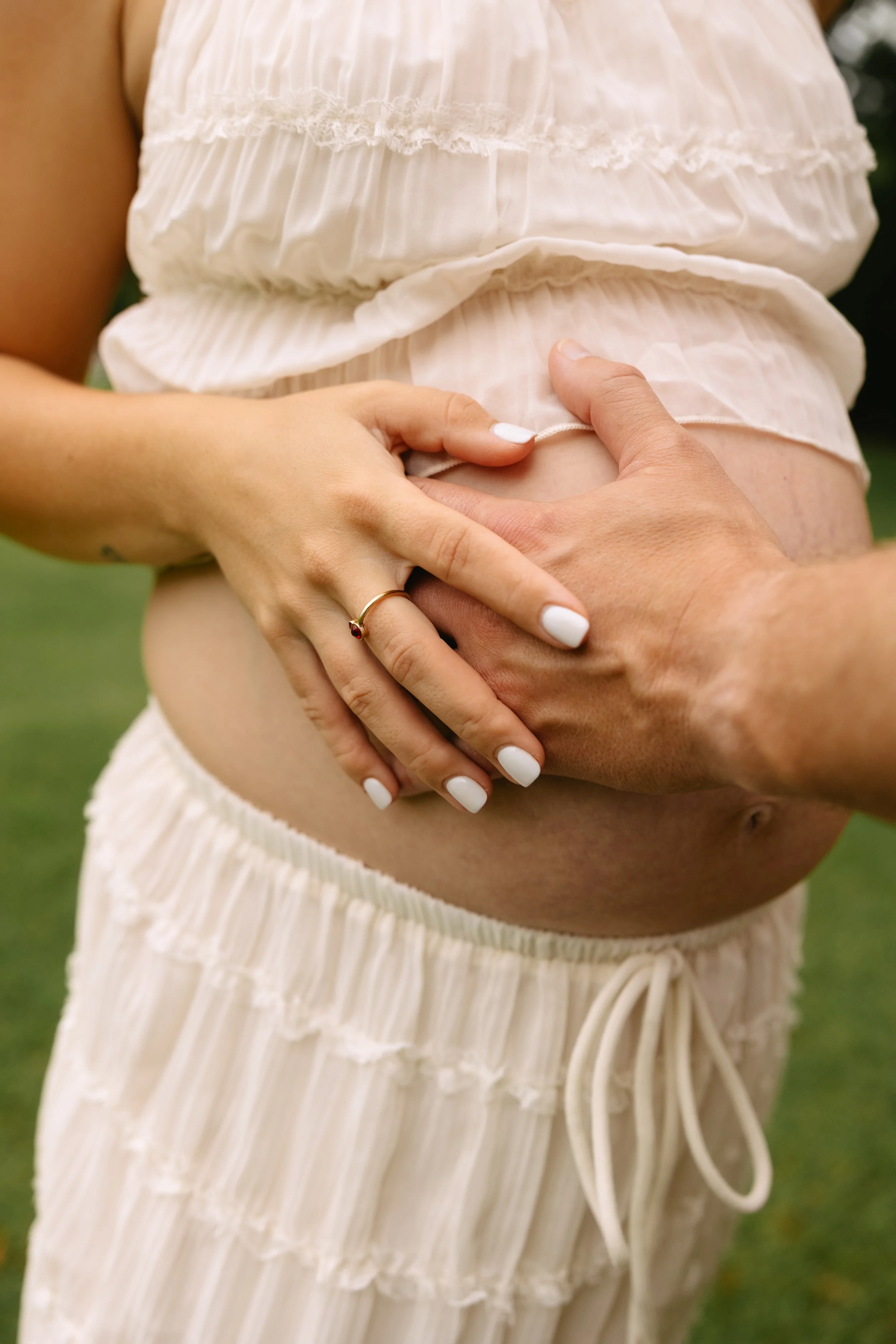 Pregnant woman wearing a white, ruffled, loose dress with her hands on her baby bump, one hand wearing a gold ring with a red gemstone, outdoors on a green lawn.