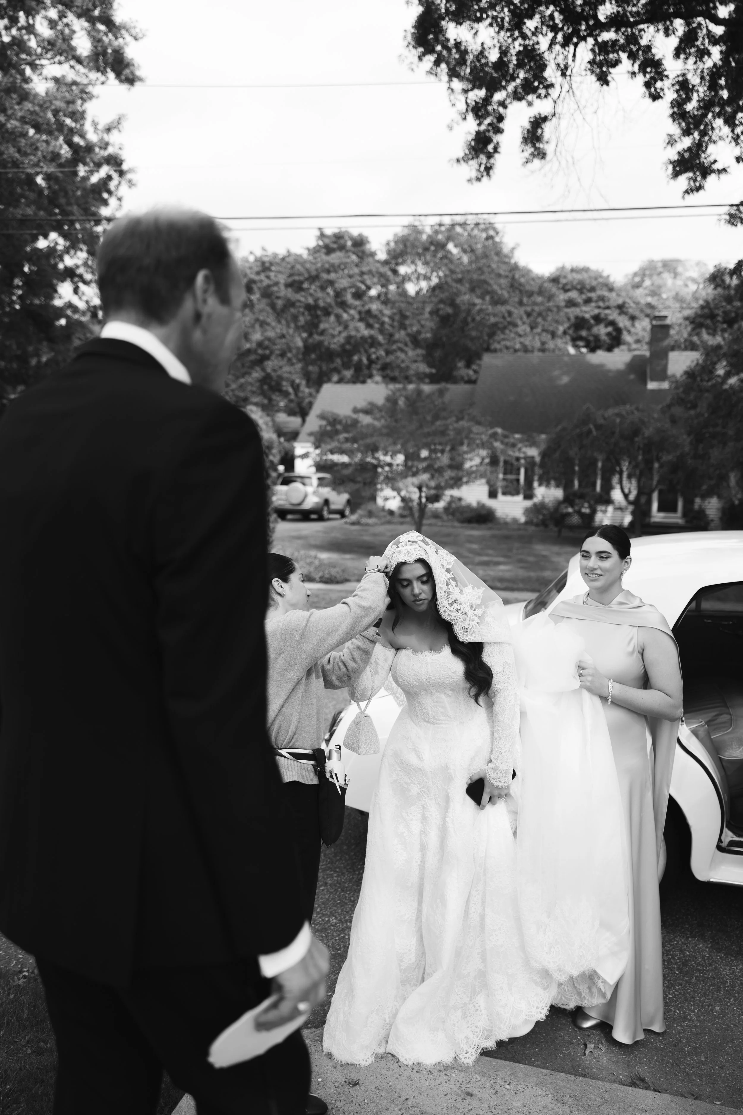 Black and white photo of a bride getting ready for her wedding, assisted by a woman placing a veil on her head, while an officiant or another woman looks on smiling, with a car and trees in the background.