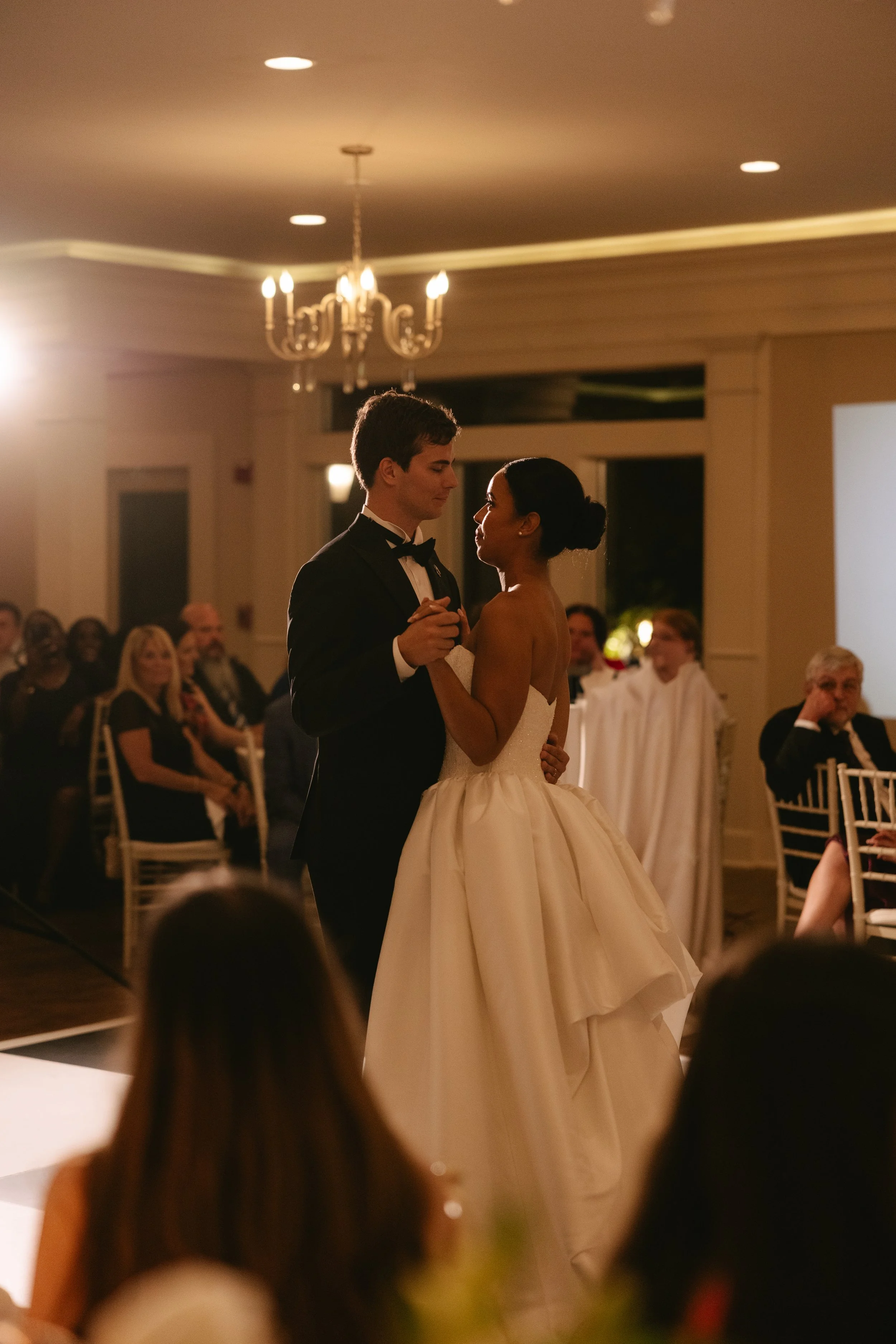 A bride and groom dance together at their wedding reception, with guests watching in the background.