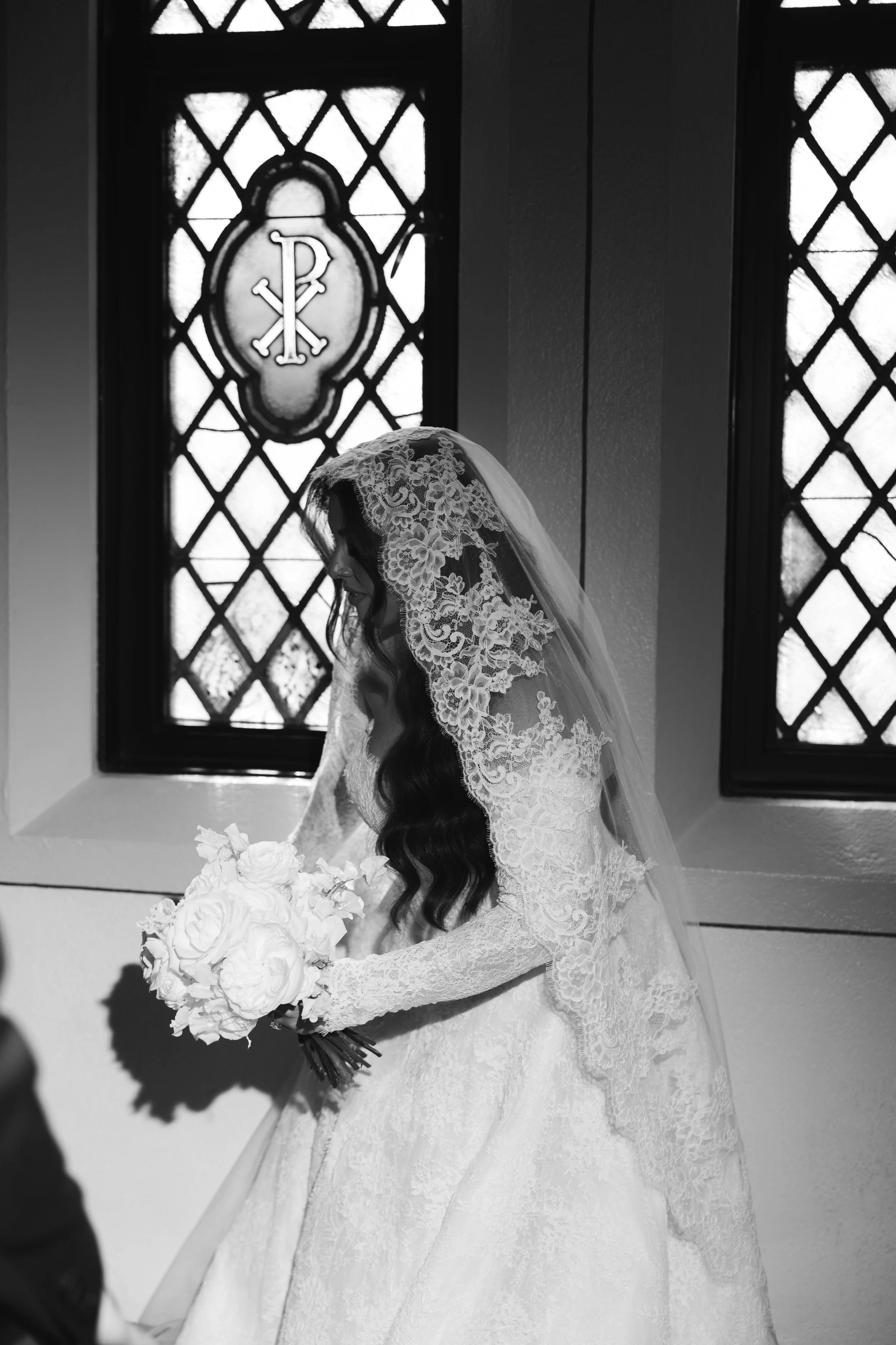 A bride in a lace wedding dress and veil holding a bouquet of white roses, standing inside a chapel with stained glass windows behind her.